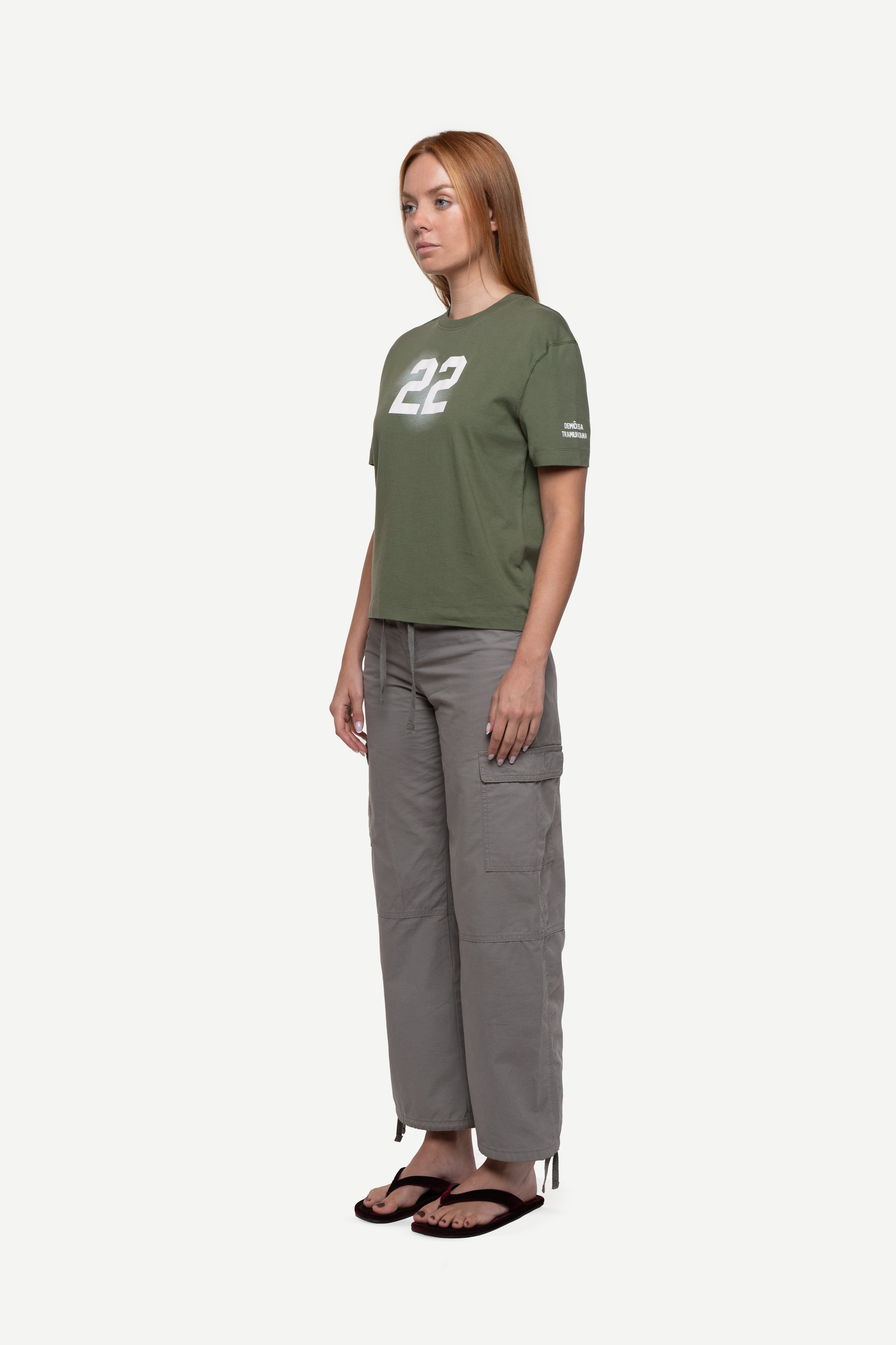 side view of woman wearing a regular fit green short-sleeved t-shirt with ivory cloudy "22" print on the chest and "demôssa, tramuntana" sleeve print, grey cargo pants and flip flops on a white background
