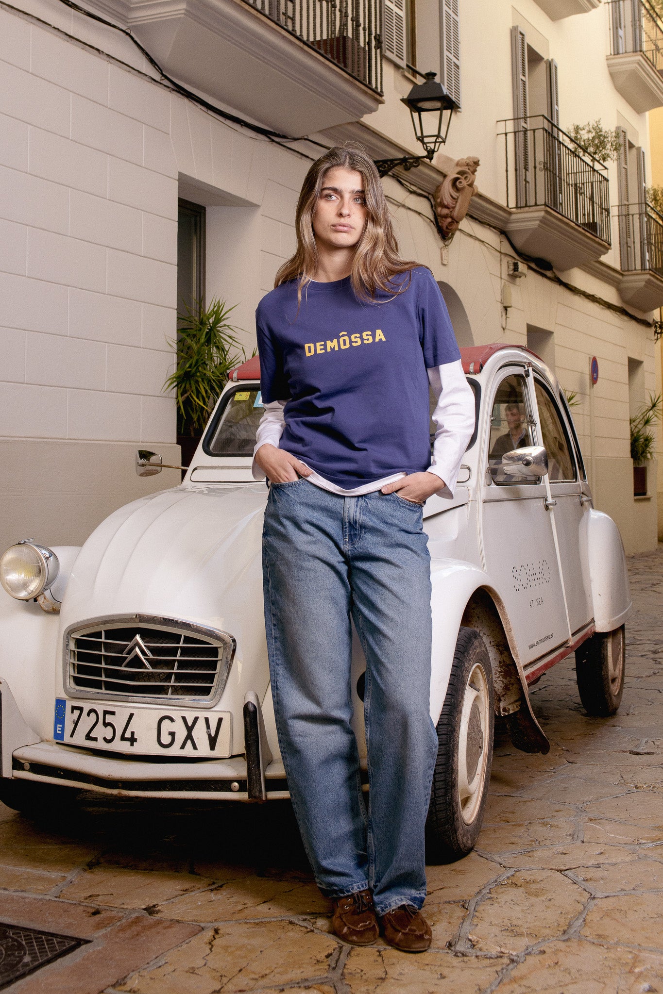 Girl wearing a blue shirt with 'Demôssa' logo standing next to a vintage car on a street.