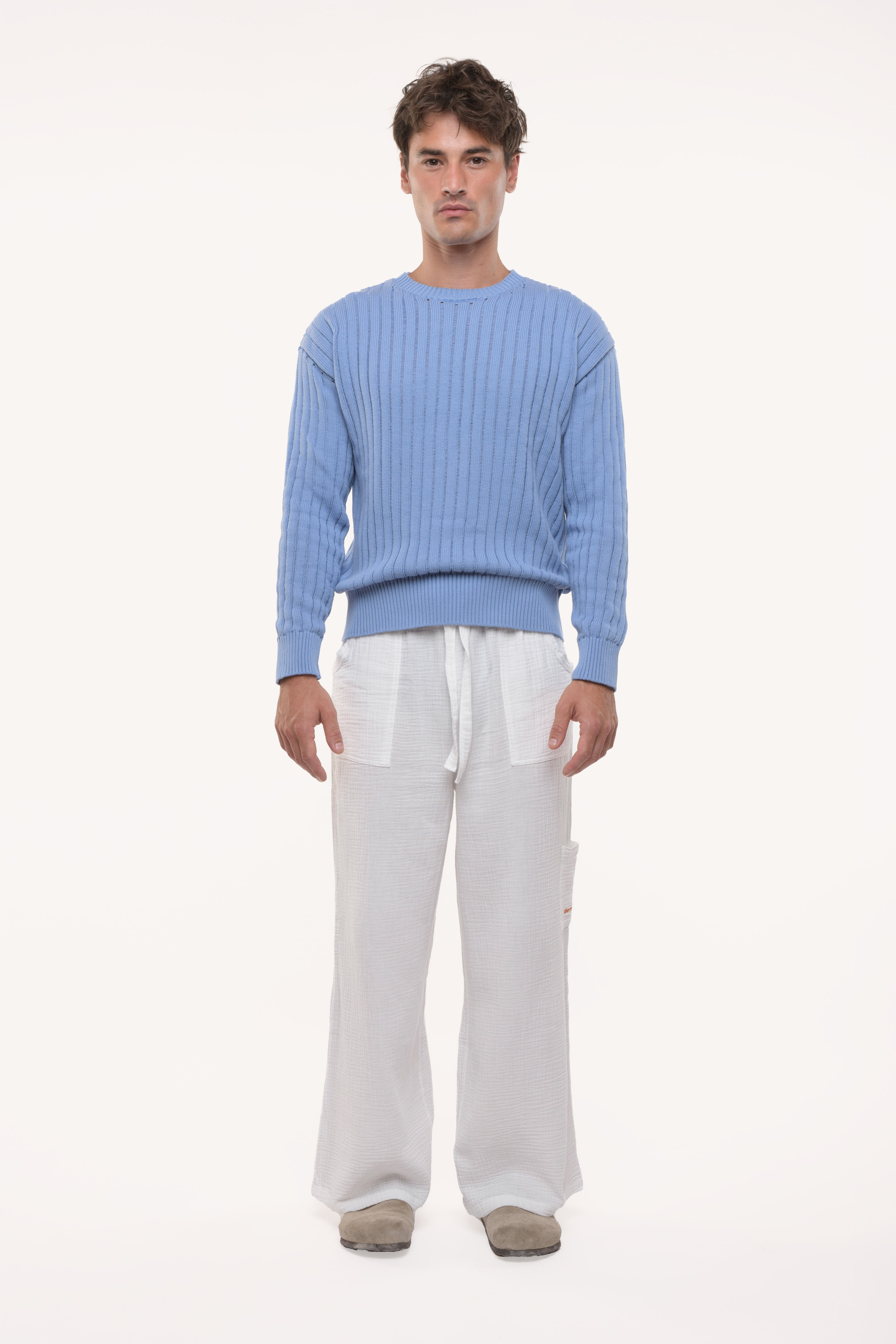 Man wearing a blue open knit sweater and white pants on a white background