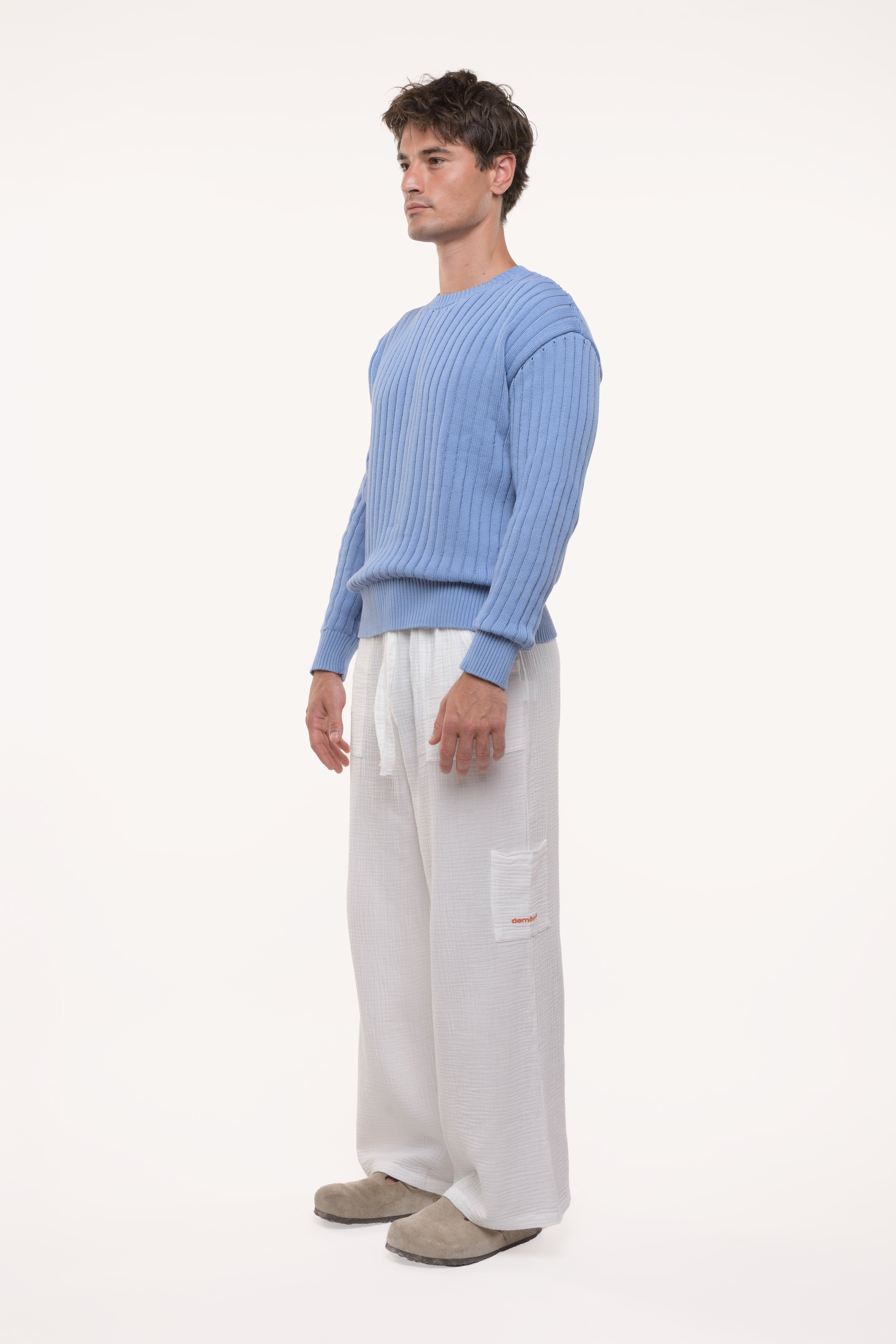 Side view of a man wearing a blue ribbed sweater and white pants on a white background