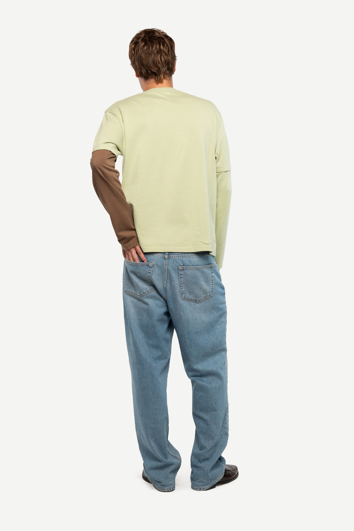 A man viewed from behind wearing a light sage green long-sleeve shirt with brown sleeves, paired with blue jeans and dark shoes, against a plain light gray background.