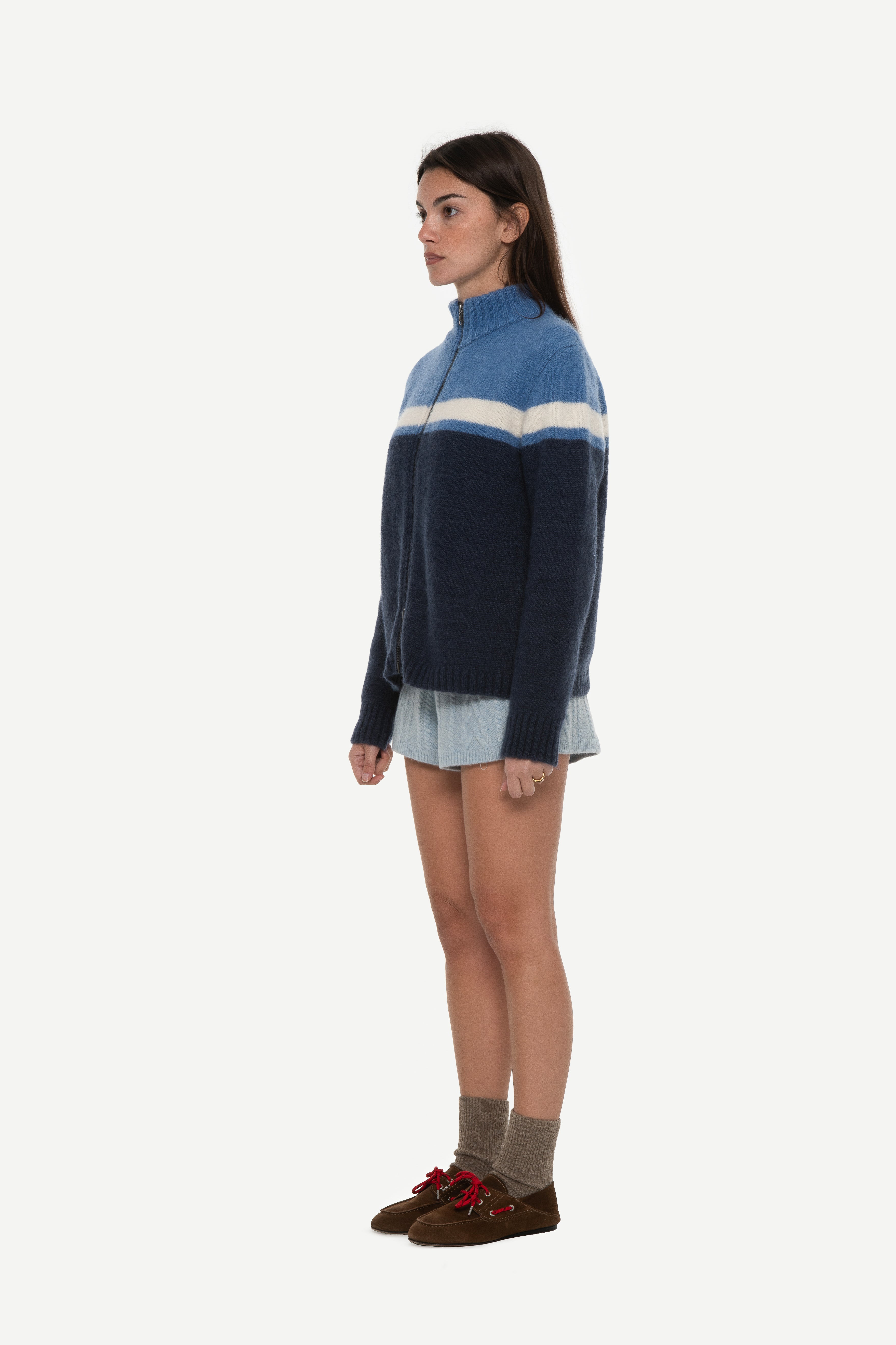 side view of woman wearing a mohair zip cardigan with two shades of blue and white stripe at chest and blue shorts on a white background