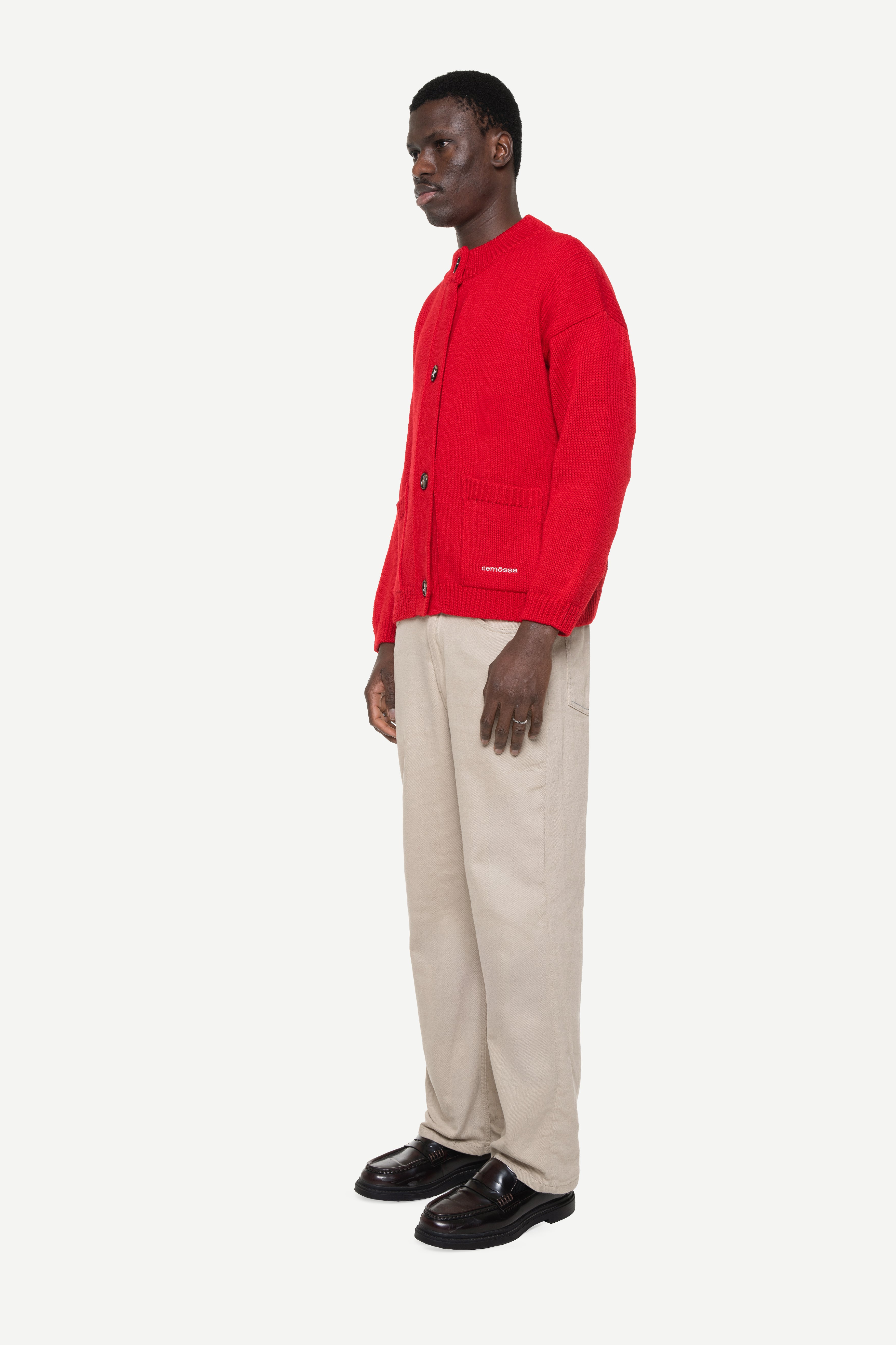 Side view of a man wearing a red buttoned cardigan with two front pockets and beige jeans on a white background