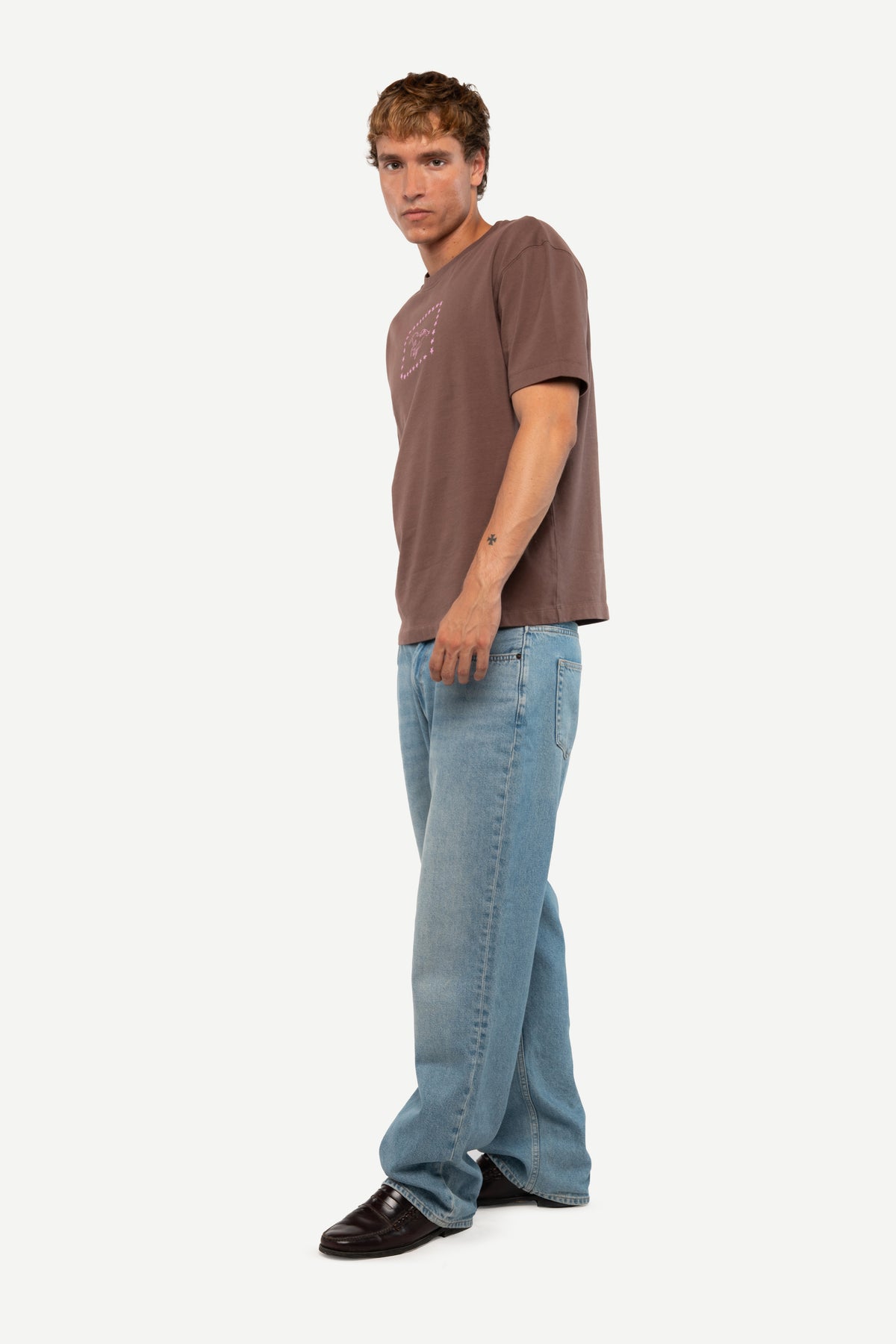 A man modeling a brown short-sleeve t-shirt and light blue wide-leg jeans against a plain light gray background.