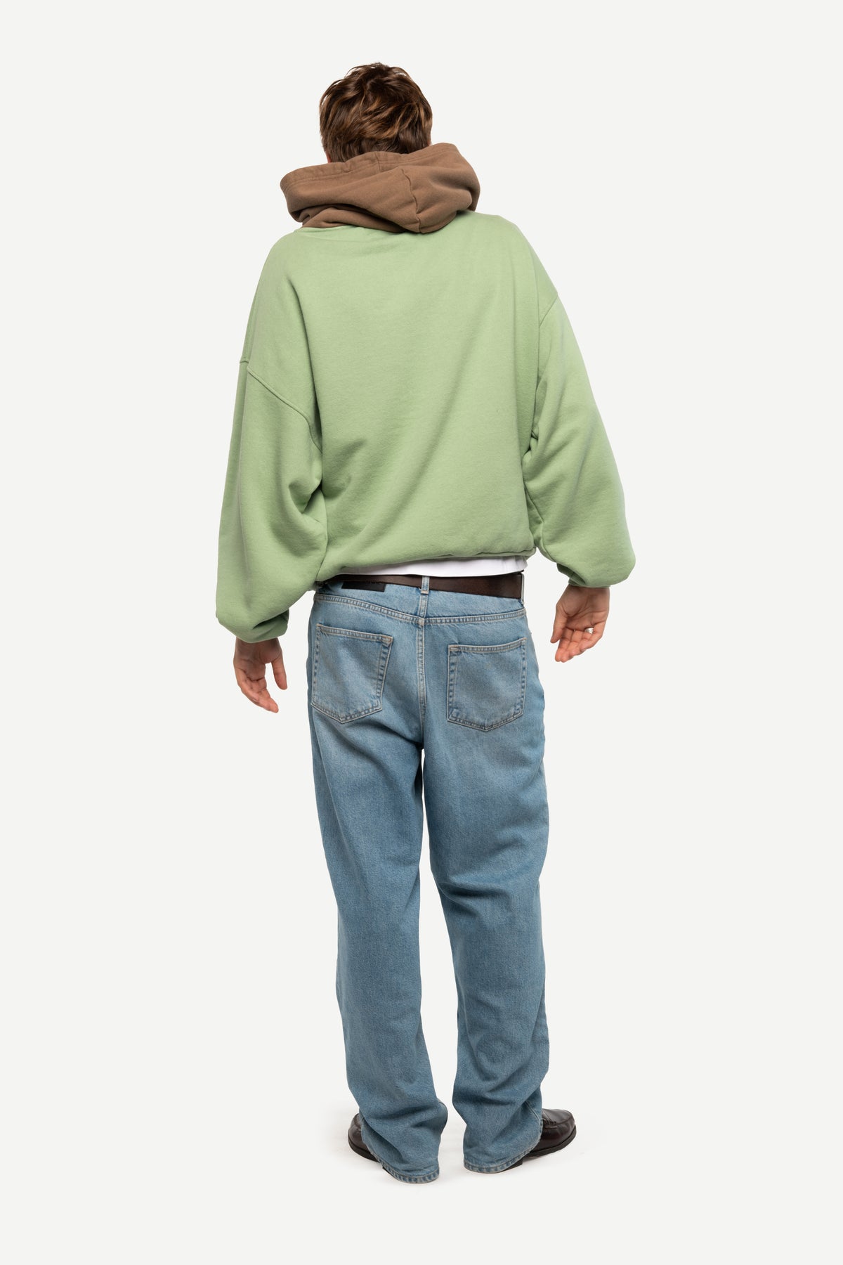 A person shown from the back wearing an oversized sage green sweatshirt with a brown hood, blue jeans, and dark shoes, photographed against a plain light gray studio background.