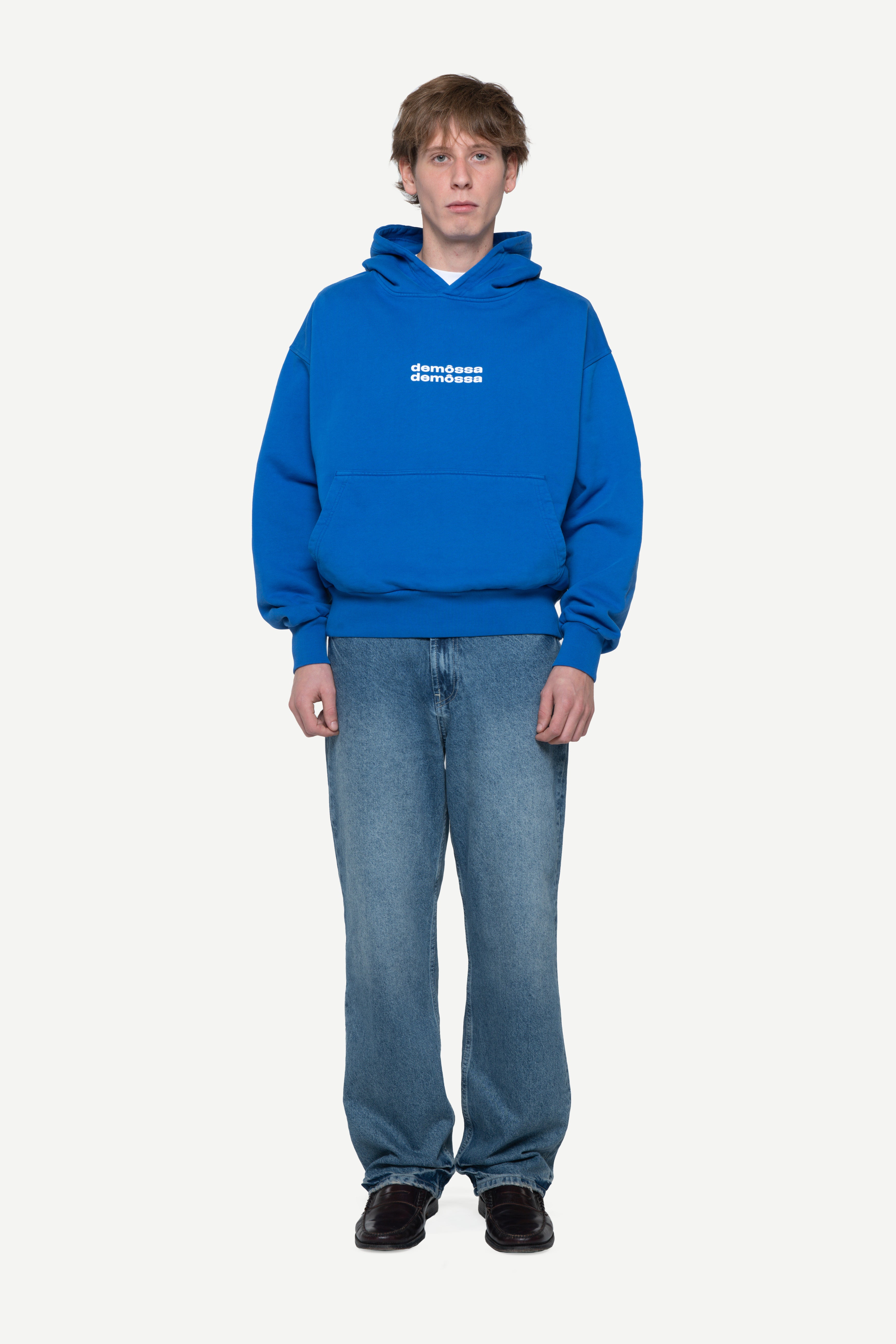 Man wearing a blue hoodie with two white demossa logos on a light gray background