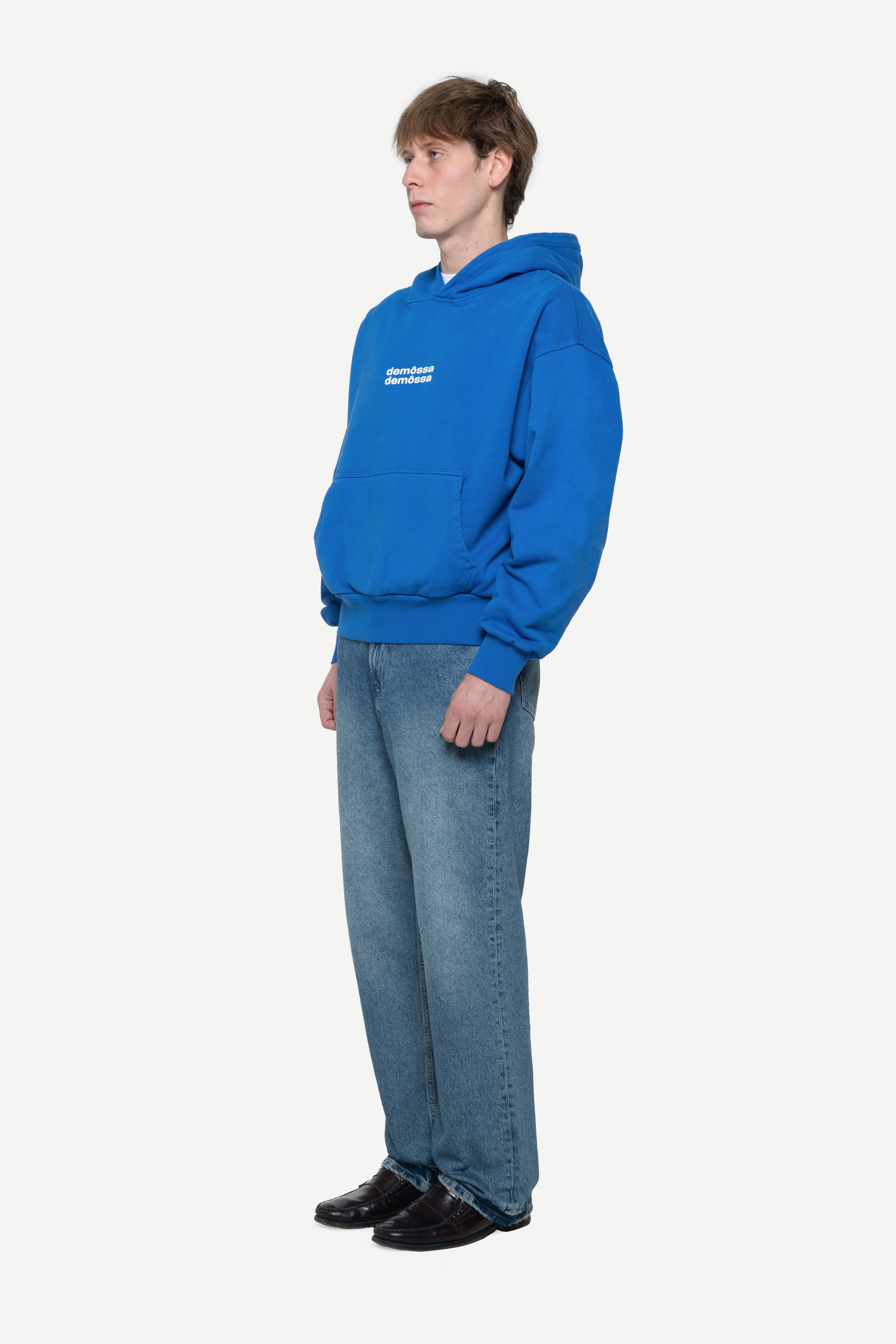 Side view of a man wearing a blue hoodie with two white demossa logos on a light gray background