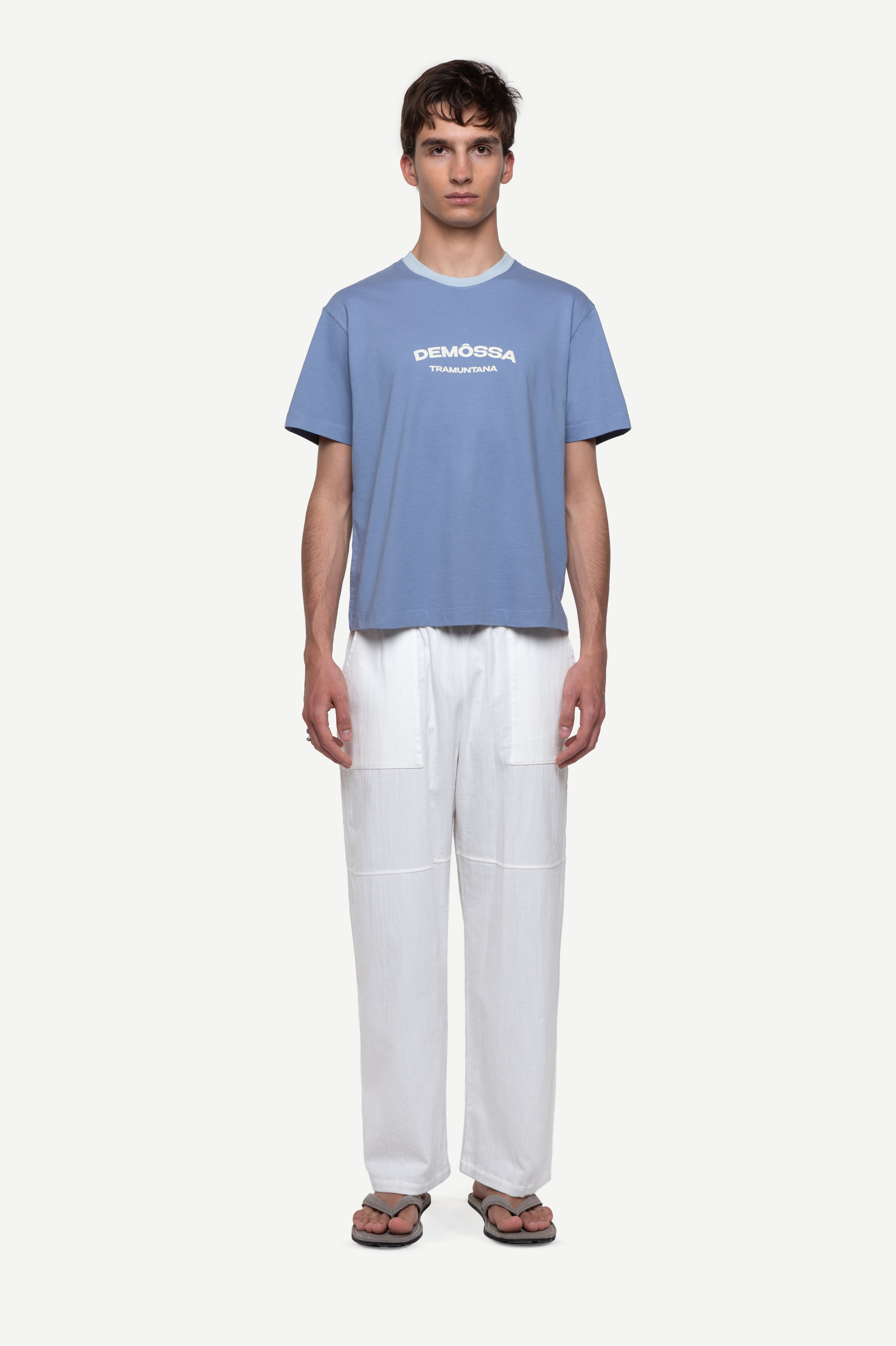 front view of man wearing a regular fit blue short-sleeved t-shirt with ivory demôssa print on the chest, white summer trousers and flip flops on a white background