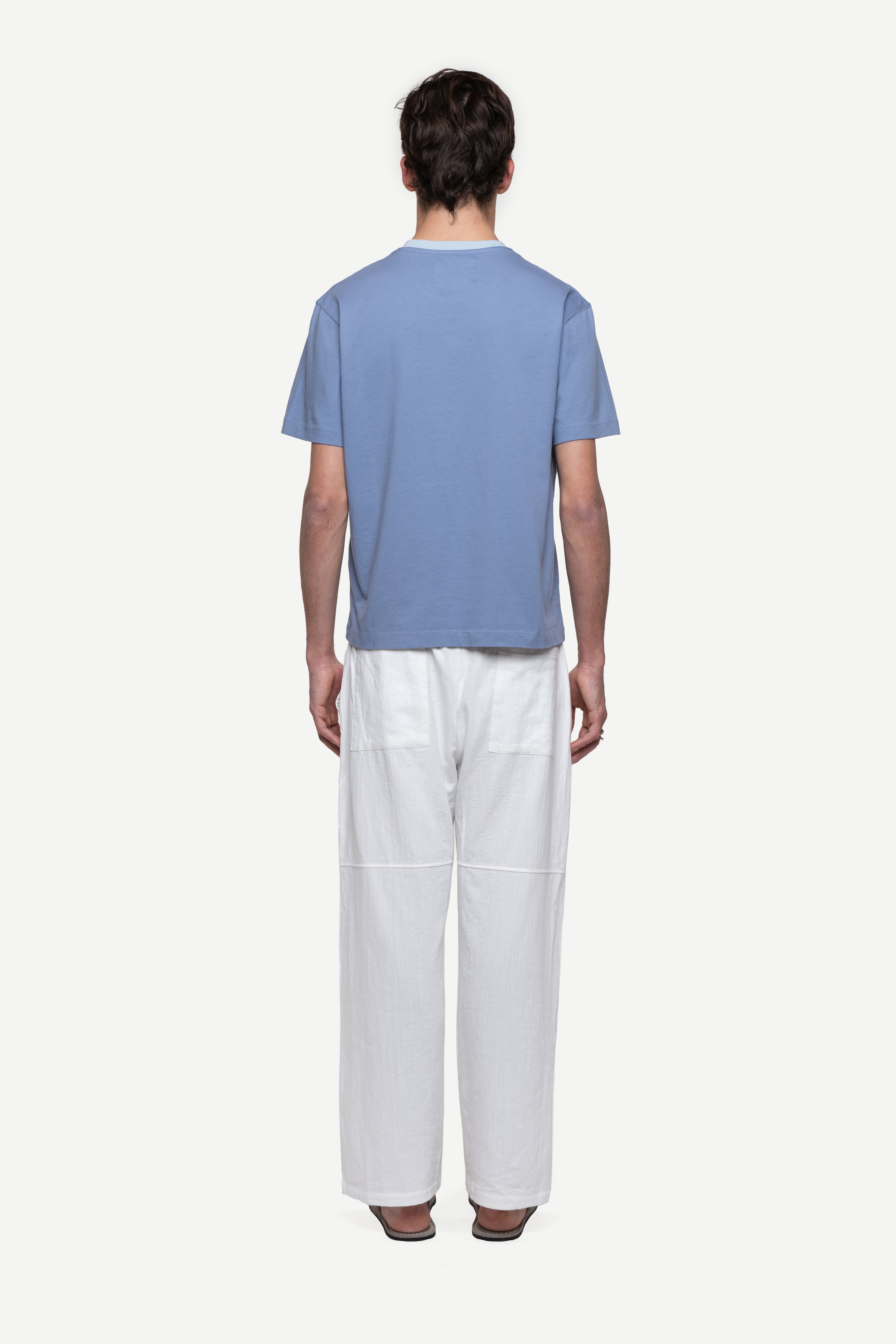 back view of man wearing a regular fit blue short-sleeved t-shirt, white summer trousers and flip flops on a white background