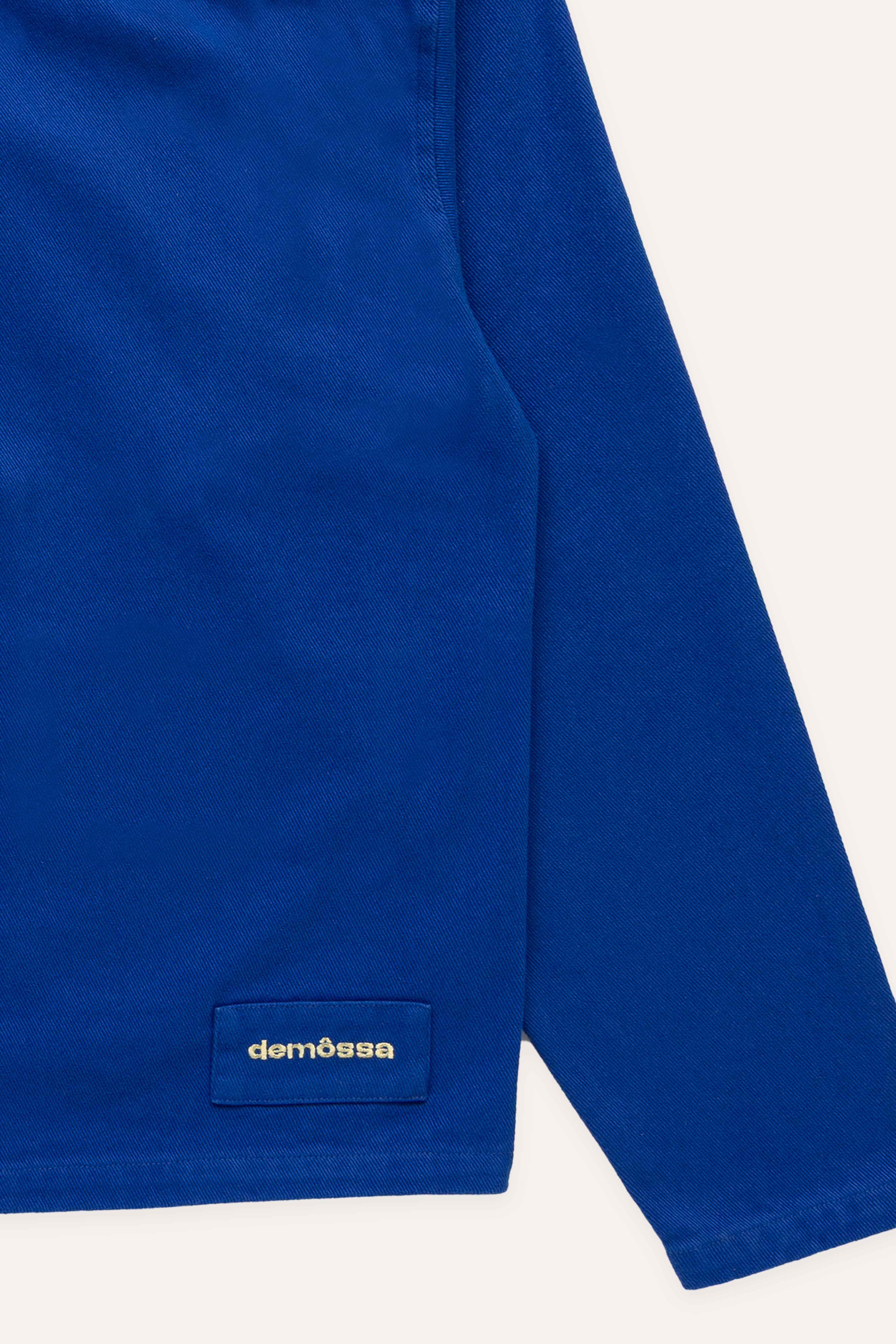 Detail of Demossa logo patch near the bottom hem on a blue overshirt