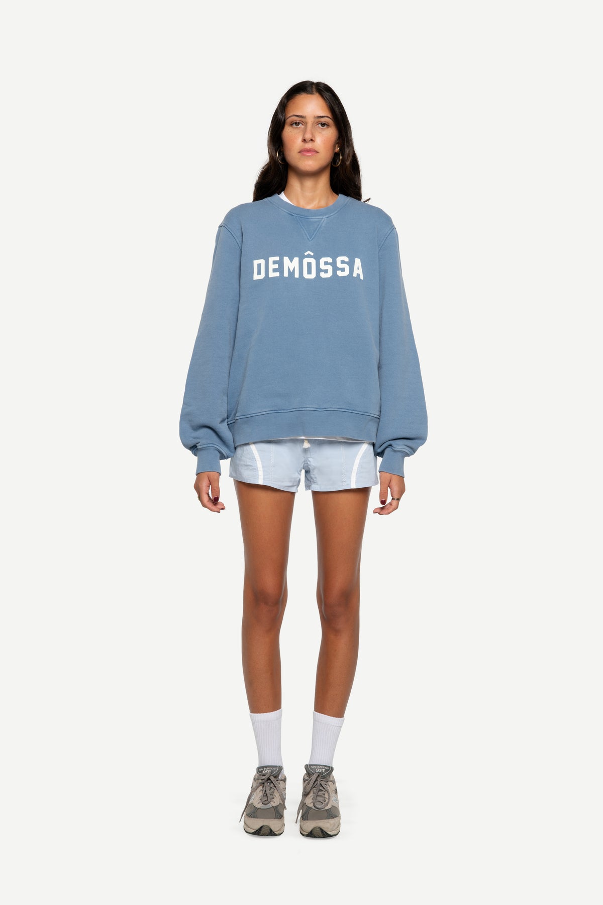 A model stands against a light gray background wearing a dusty blue crewneck sweatshirt with 'DEMÖSSA' printed in white across the chest, paired with light denim shorts, white socks, and gray sneakers.