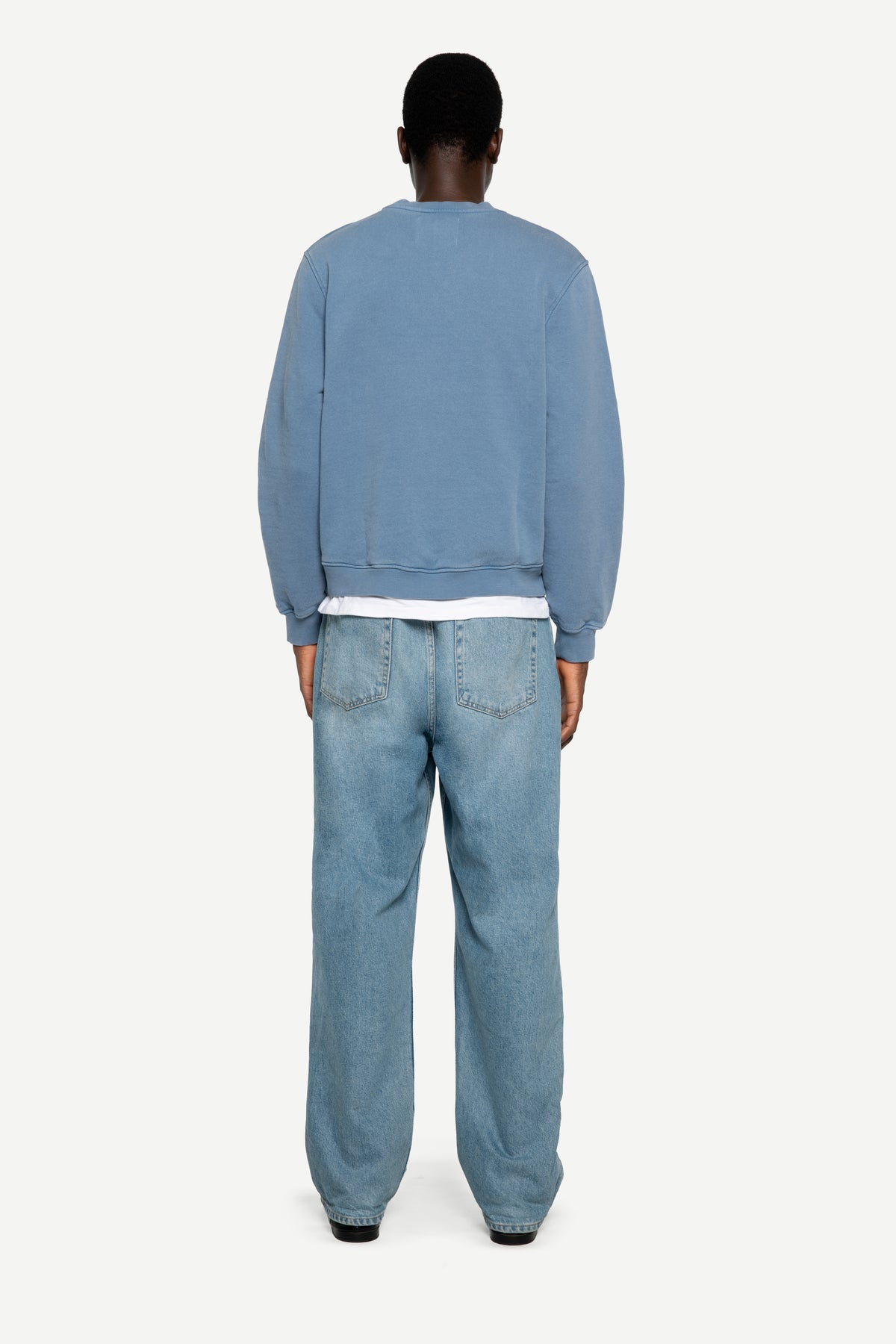 A man viewed from behind wearing a light blue crewneck sweatshirt, light-wash blue jeans, and black shoes, standing against a plain light gray studio background.