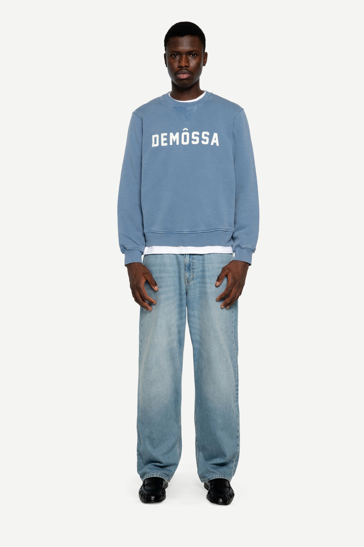 A man modeling a light blue crewneck sweatshirt with 'DEMÔSSA' printed in white across the chest, paired with light-wash jeans and black shoes, against a plain light gray background.