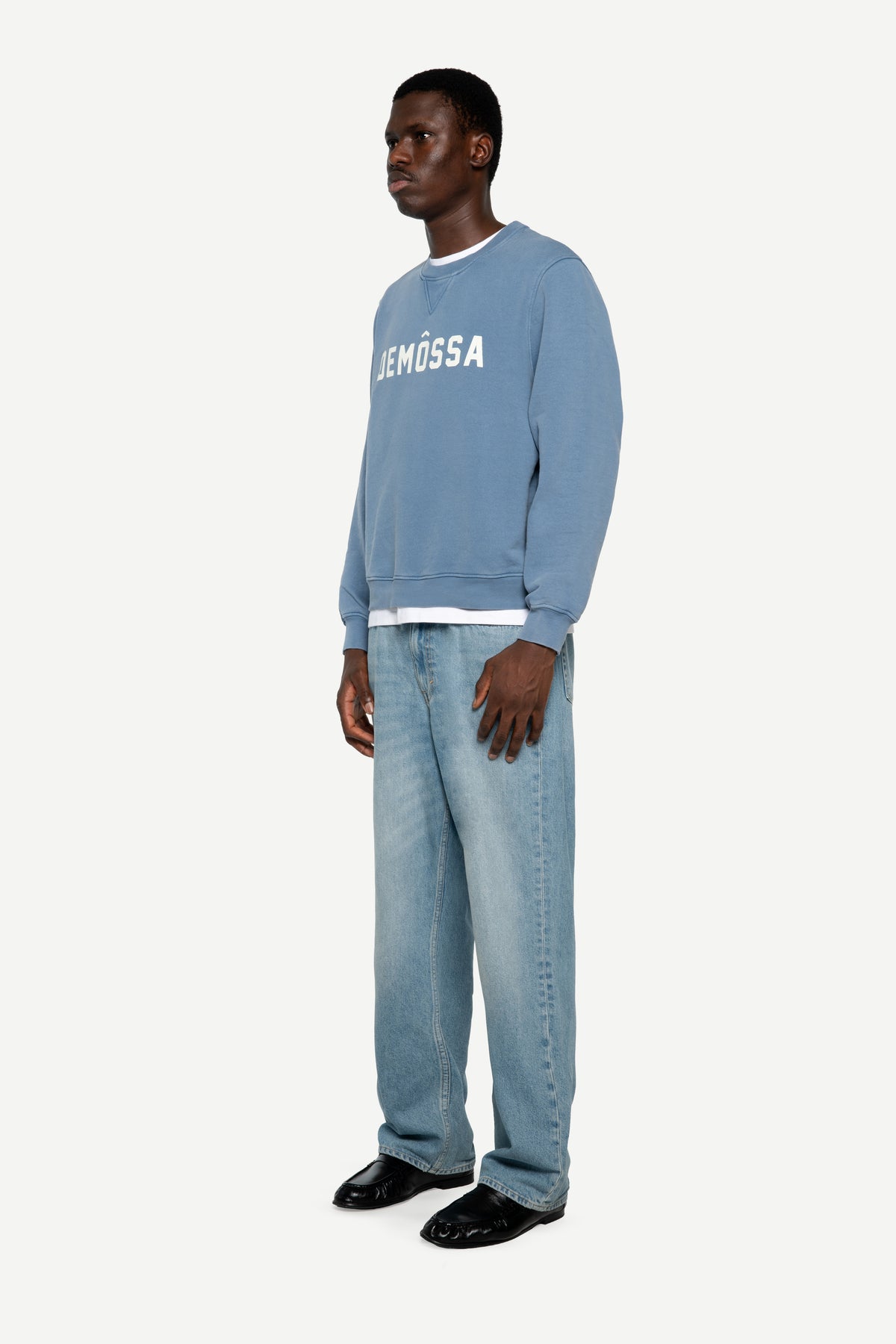 A man wearing a light blue crewneck sweatshirt with 'DEMÔSSA' printed in white across the chest, paired with light blue jeans and black shoes, standing against a plain light gray background.