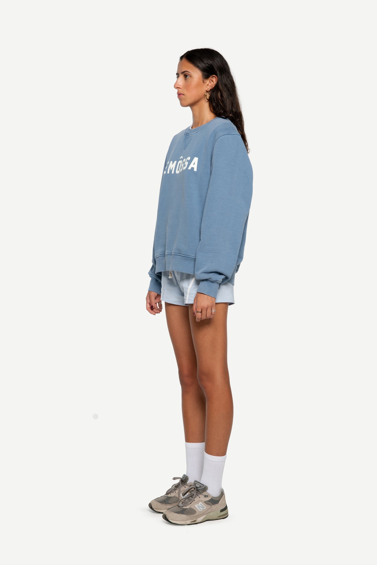 A woman stands in profile wearing a light blue oversized sweatshirt with white lettering, light denim shorts, white socks, and gray sneakers against a plain light gray background.