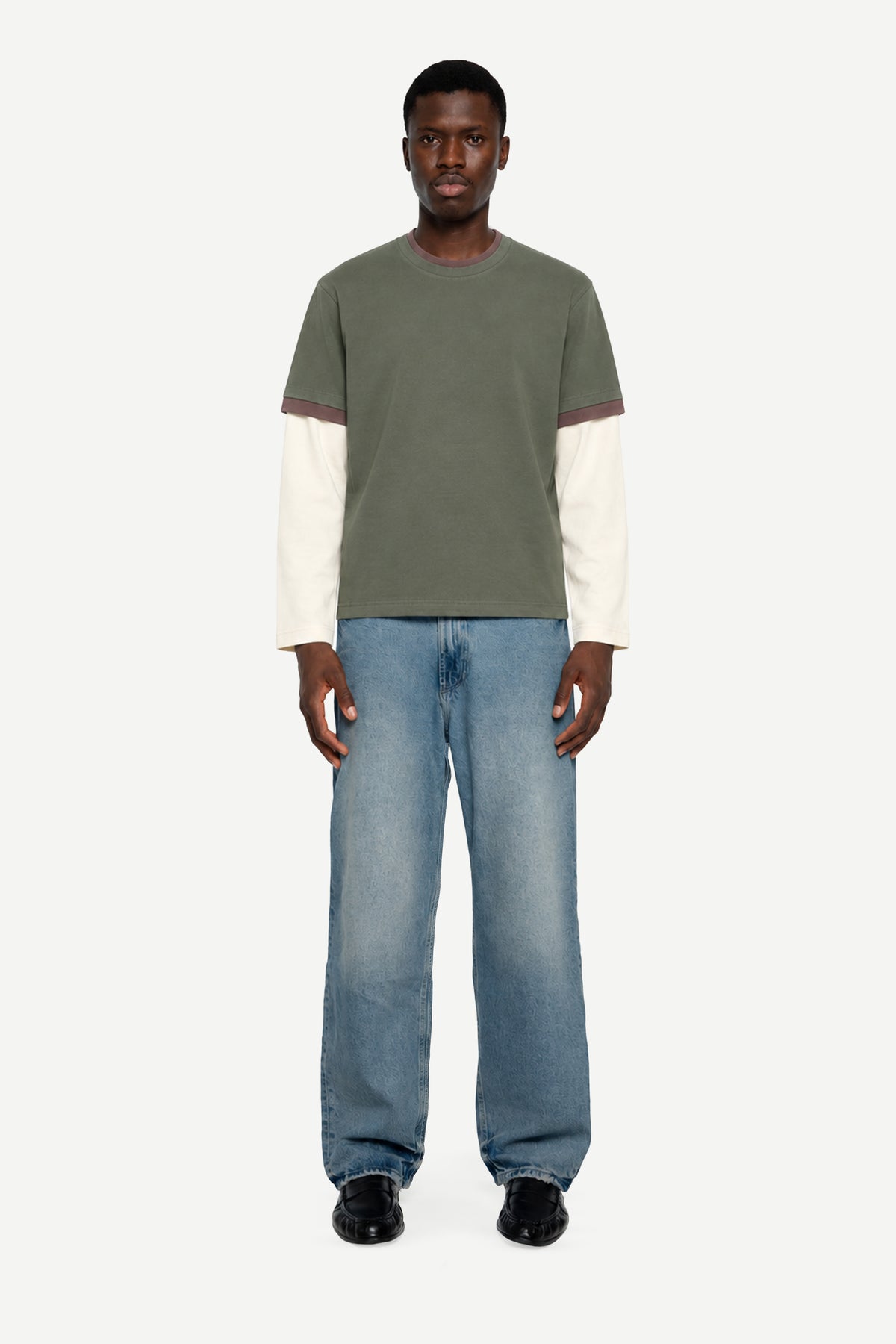 A man modeling a color-blocked knit long-sleeve shirt with an olive green body and contrasting cream sleeves, paired with light-wash blue jeans and black shoes, against a plain light gray background.