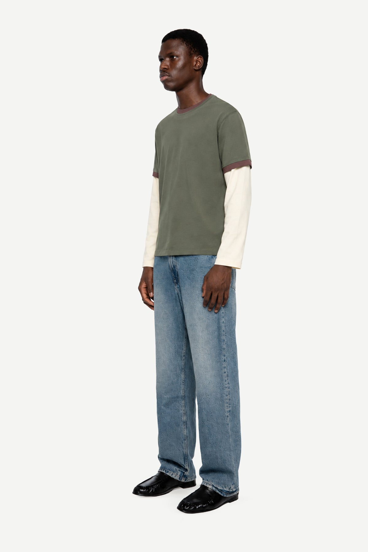 A man wearing a color-blocked short-sleeve t-shirt in olive green and cream, paired with light-wash blue jeans and black loafers, standing against a plain light gray background.