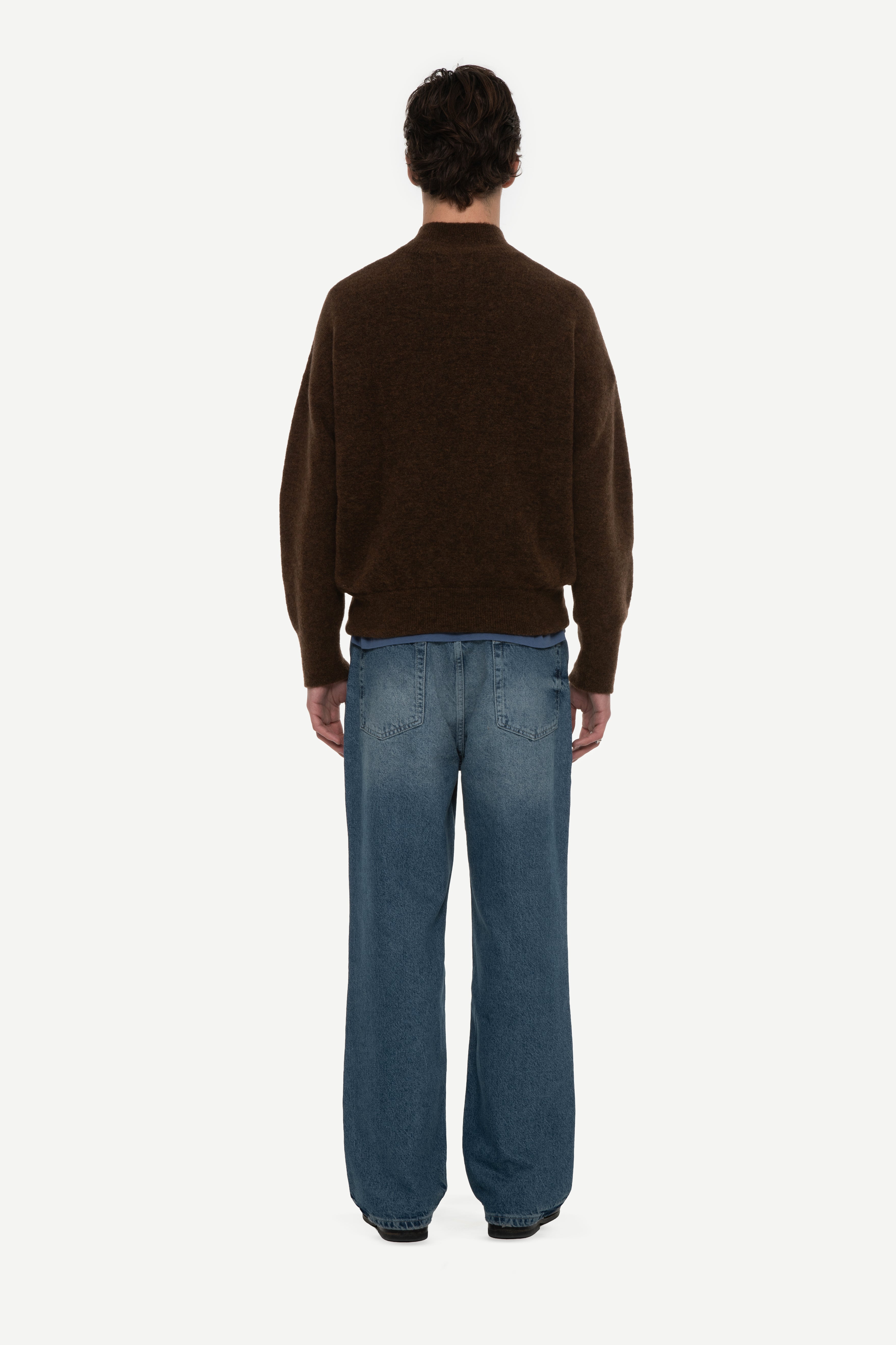 Back view of man wearing a brown mockneck sweater and blue jeans on a white background