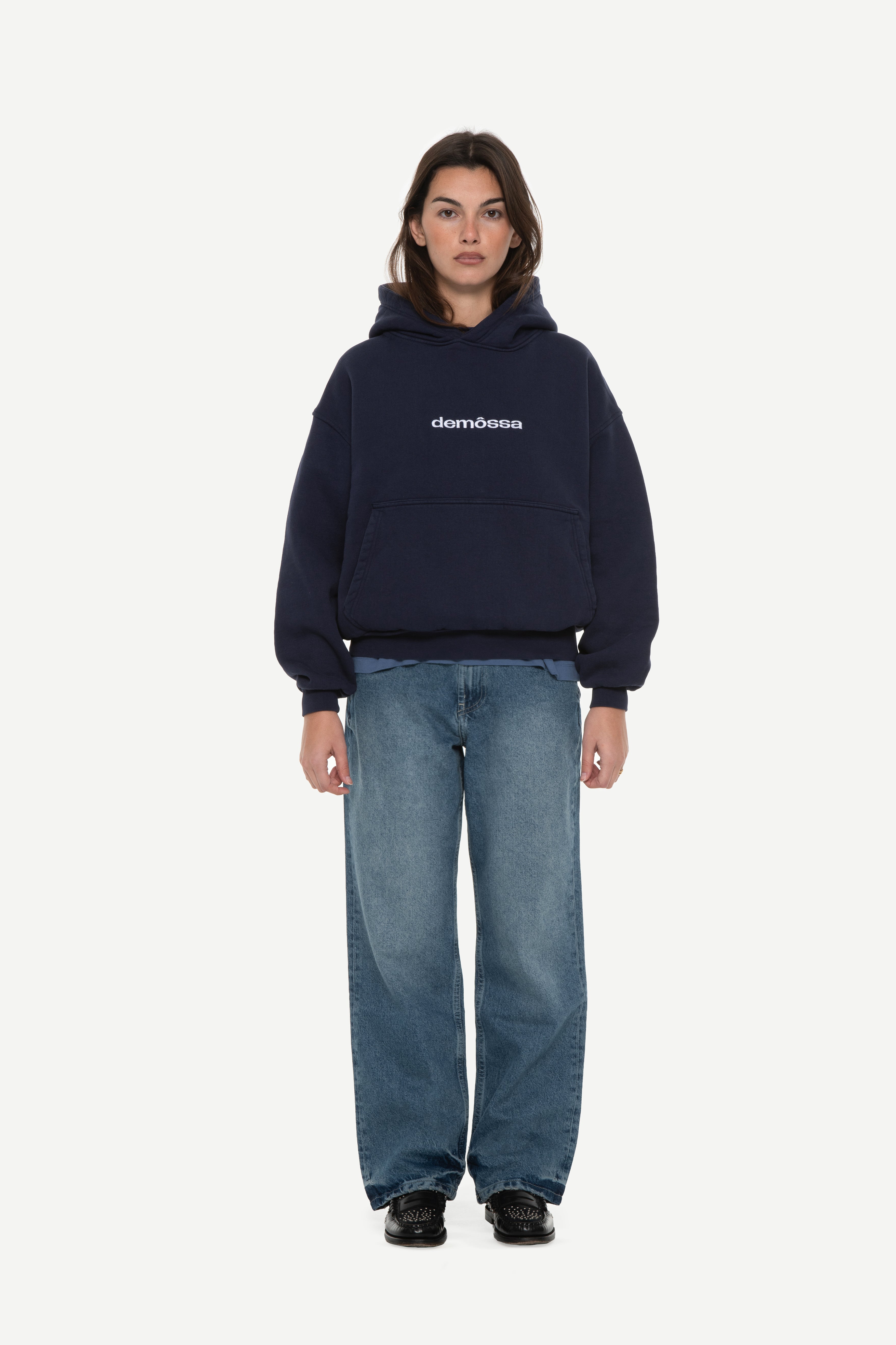front view of woman wearing a navy hoodie with “demôssa” logo embroidered on the middle chest and blue jeans on a white background