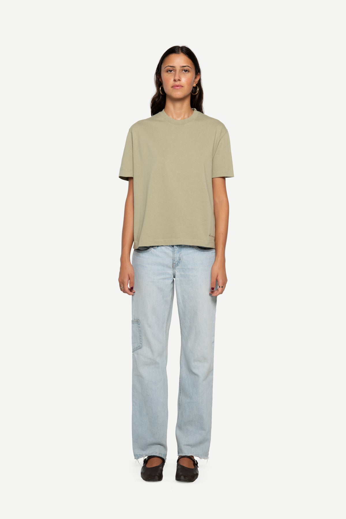 A model stands against a light gray background wearing a muted sage green short-sleeve t-shirt, light-wash blue jeans, and dark brown loafers.