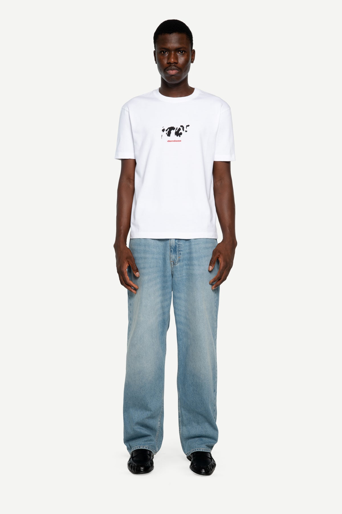 A man modeling a white short-sleeve t-shirt with a black graphic print on the chest, paired with light blue jeans and black shoes, against a plain light gray background.