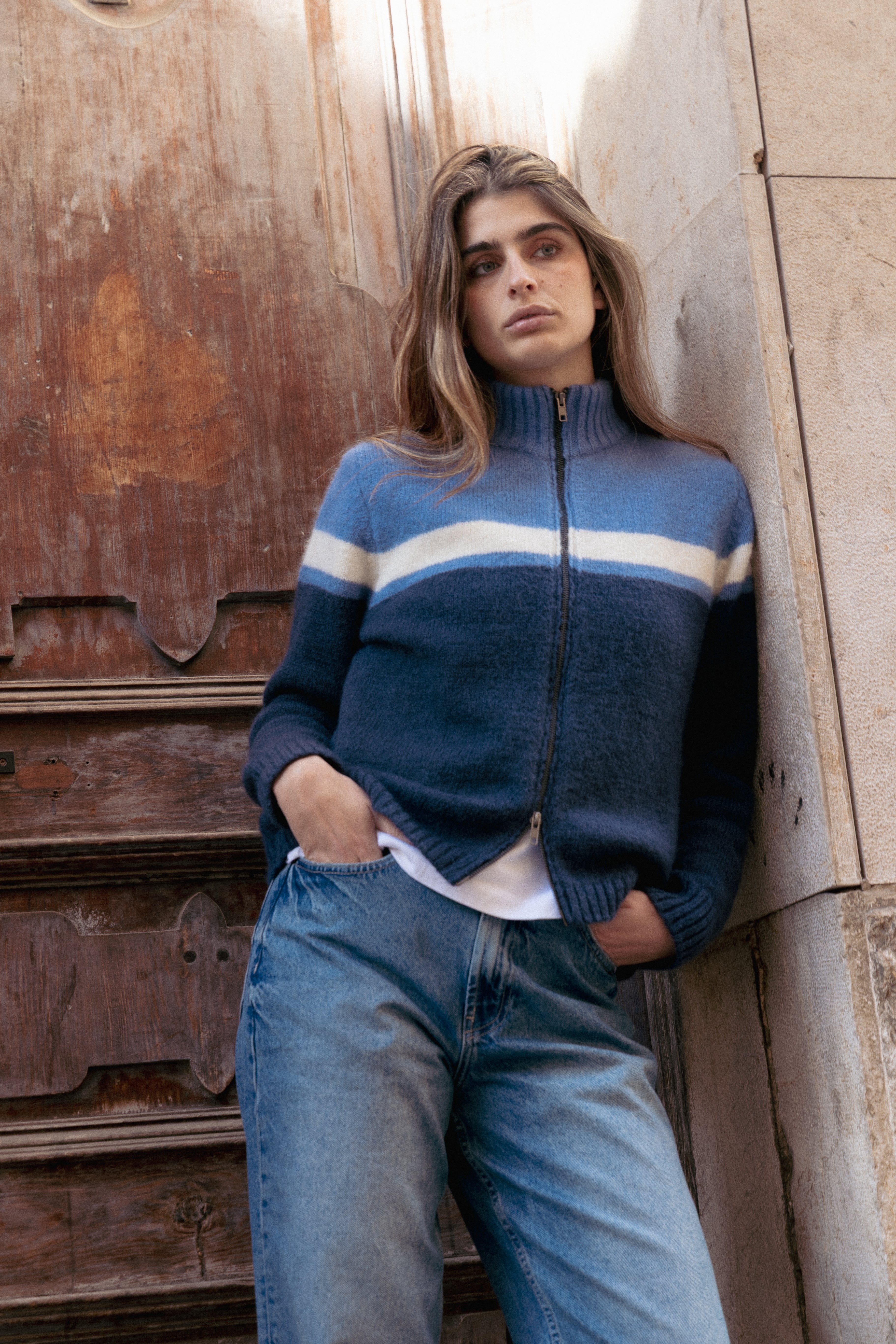 Person wearing a blue sweater with a white stripe leaning against a wood door wall.