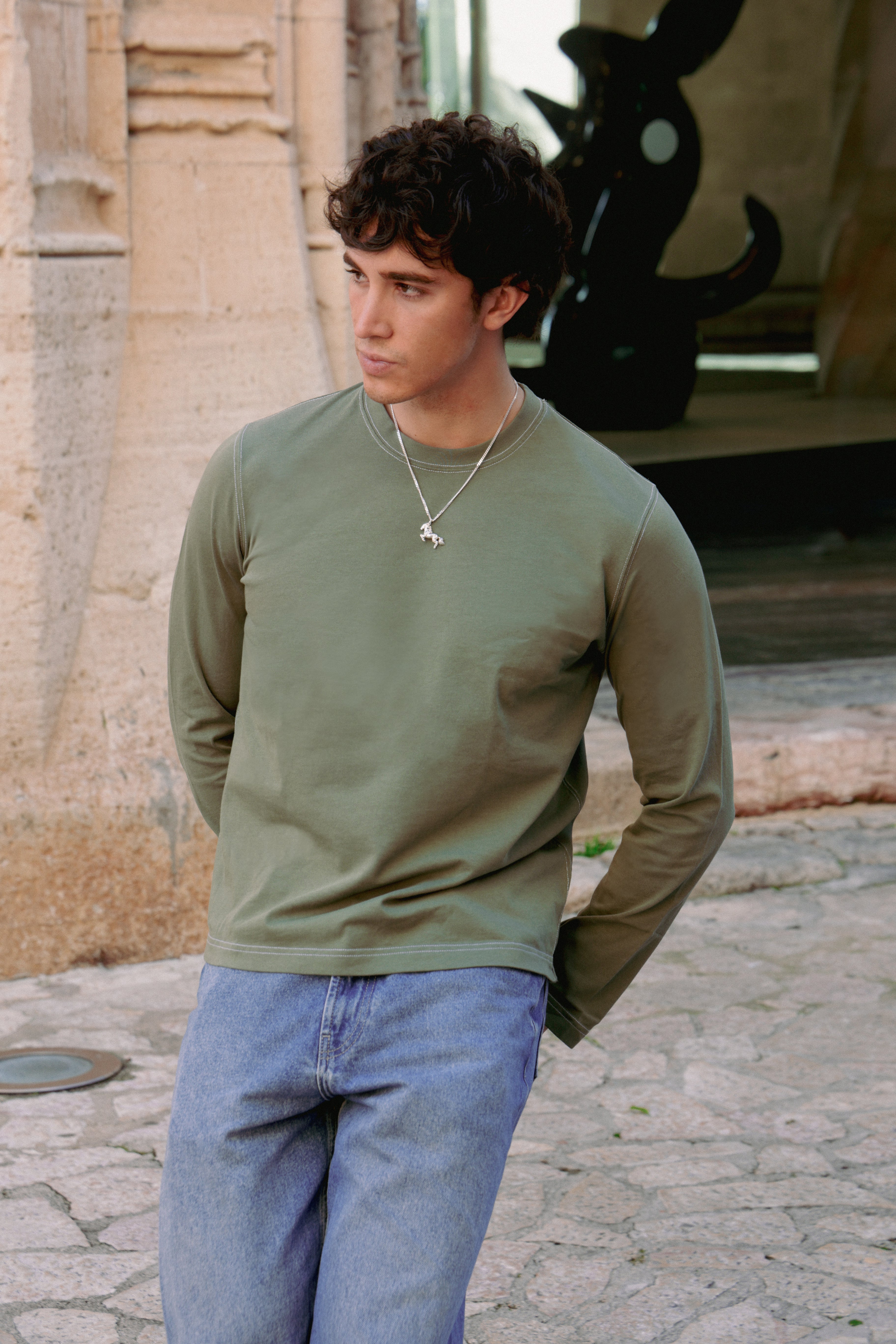 Man wearing a green long-sleeve shirt and blue jeans standing outdoors.