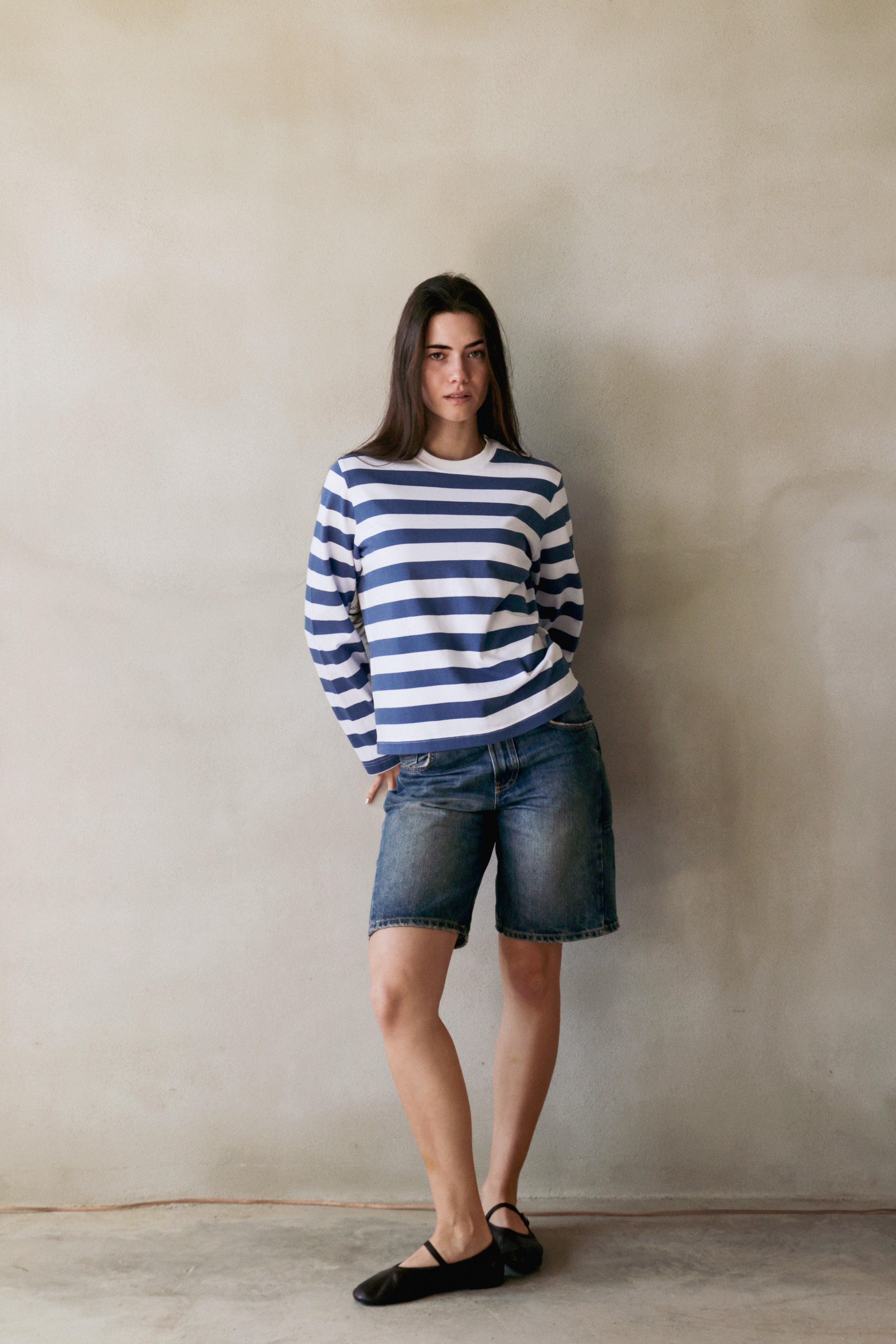 Woman wearing a striped longsleeve t-shirt and denim shorts against a plain wall.