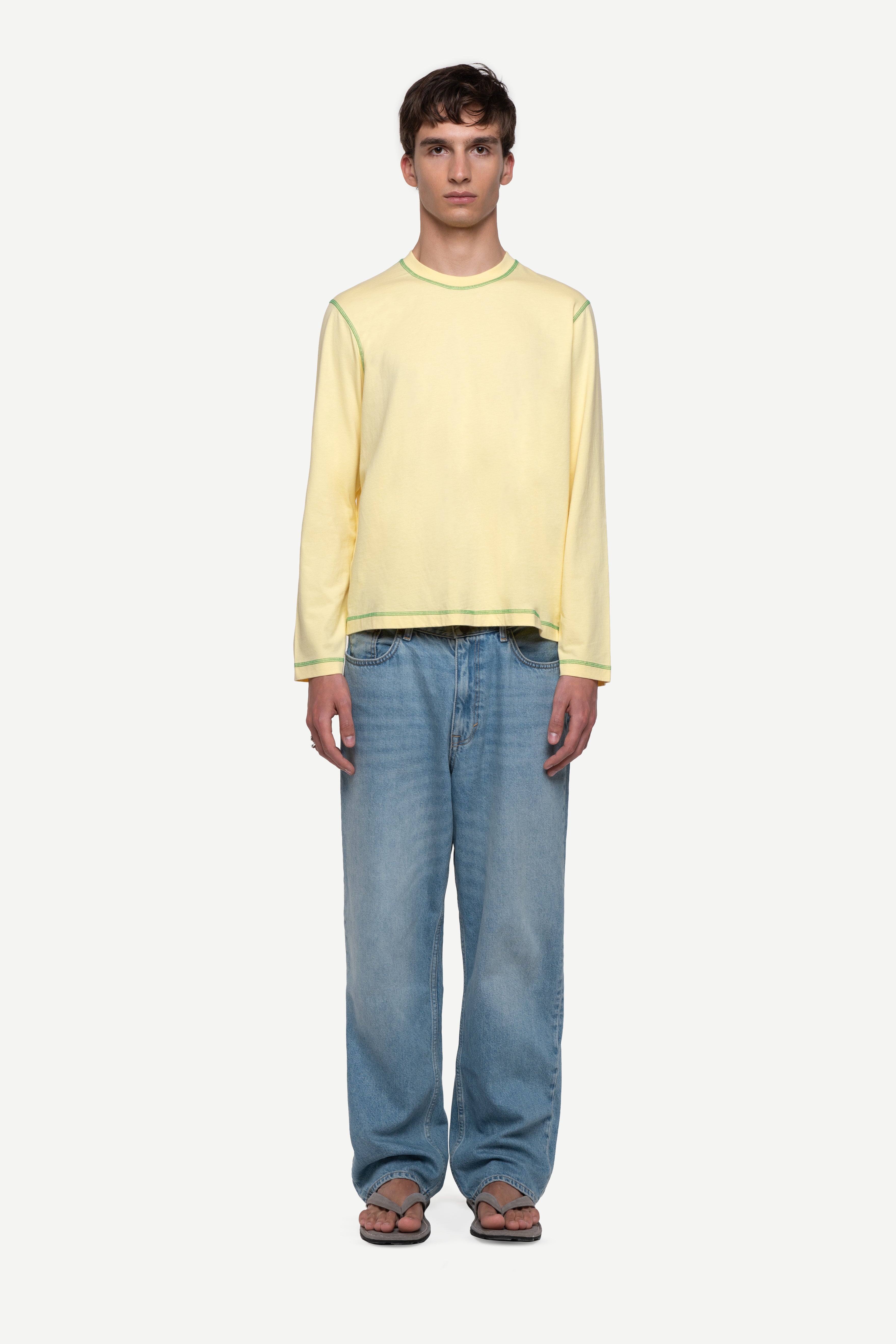 front view of man wearing a regular fit pale yellow red long-sleeved t-shirt with contrast green over-lock stitching, blue jeans and flip flops on a white background