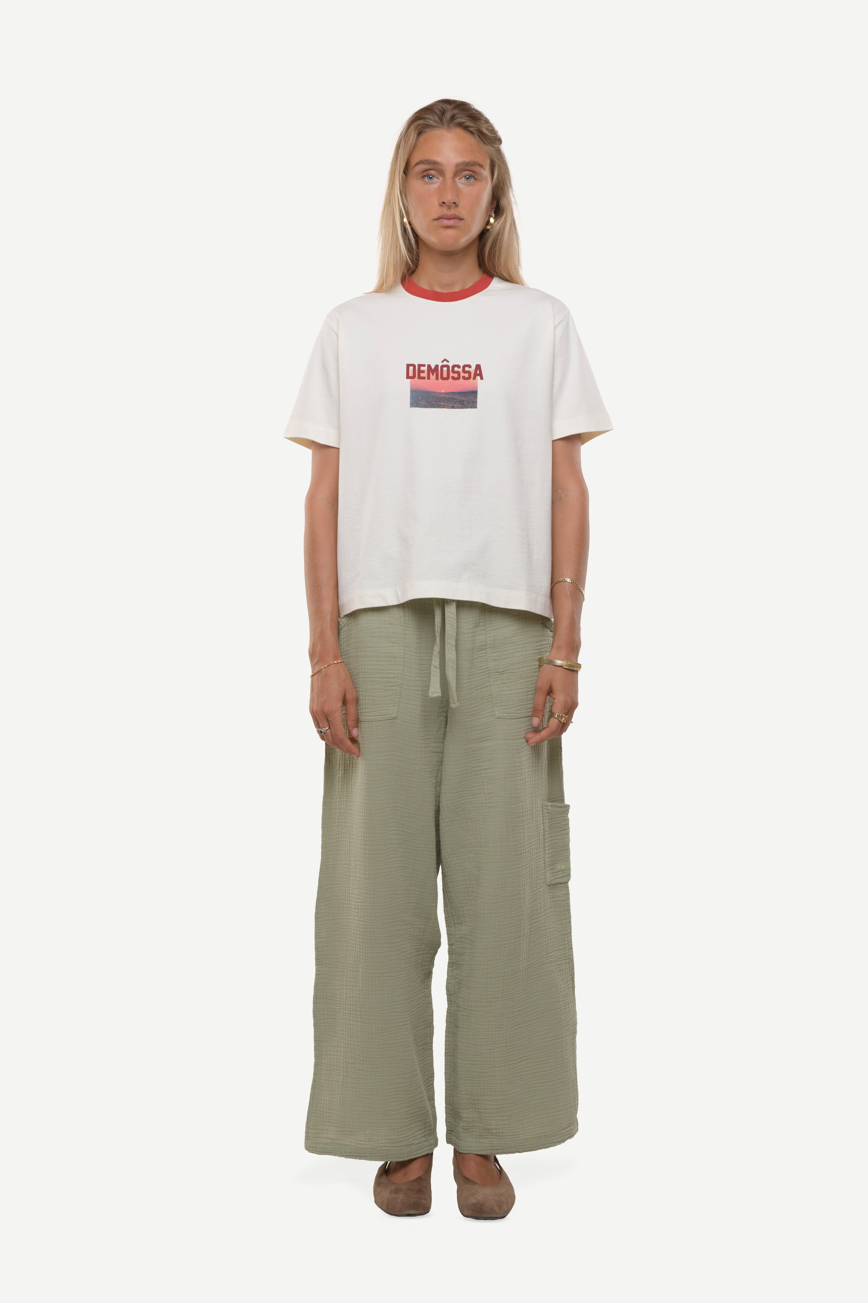 woman wearing a white t-shirt with red demossa logo and sunset picture with green cotton pants white background
