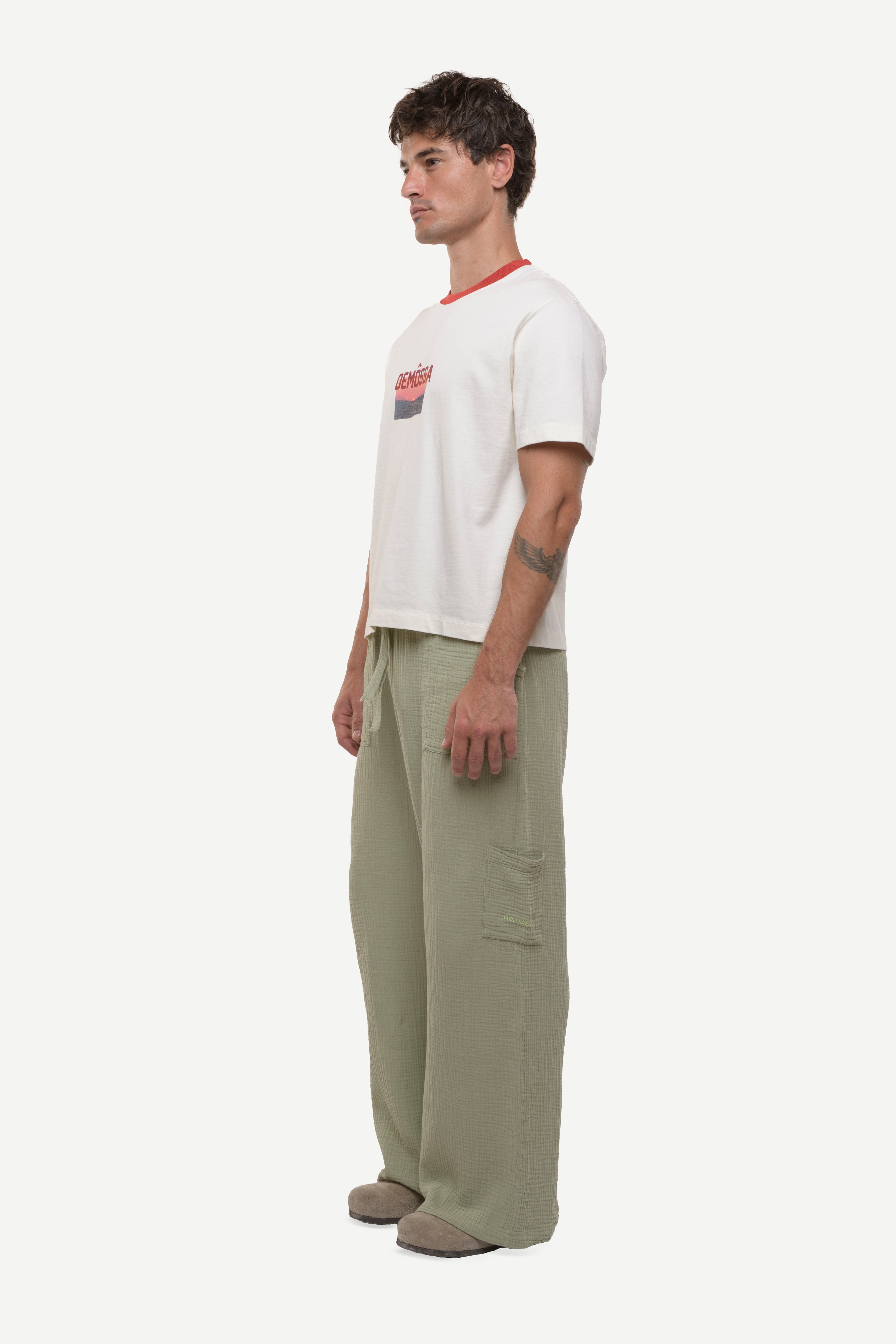 Side view of man wearing a white t-shirt with red demossa logo and sunset picture with green cotton pants white background