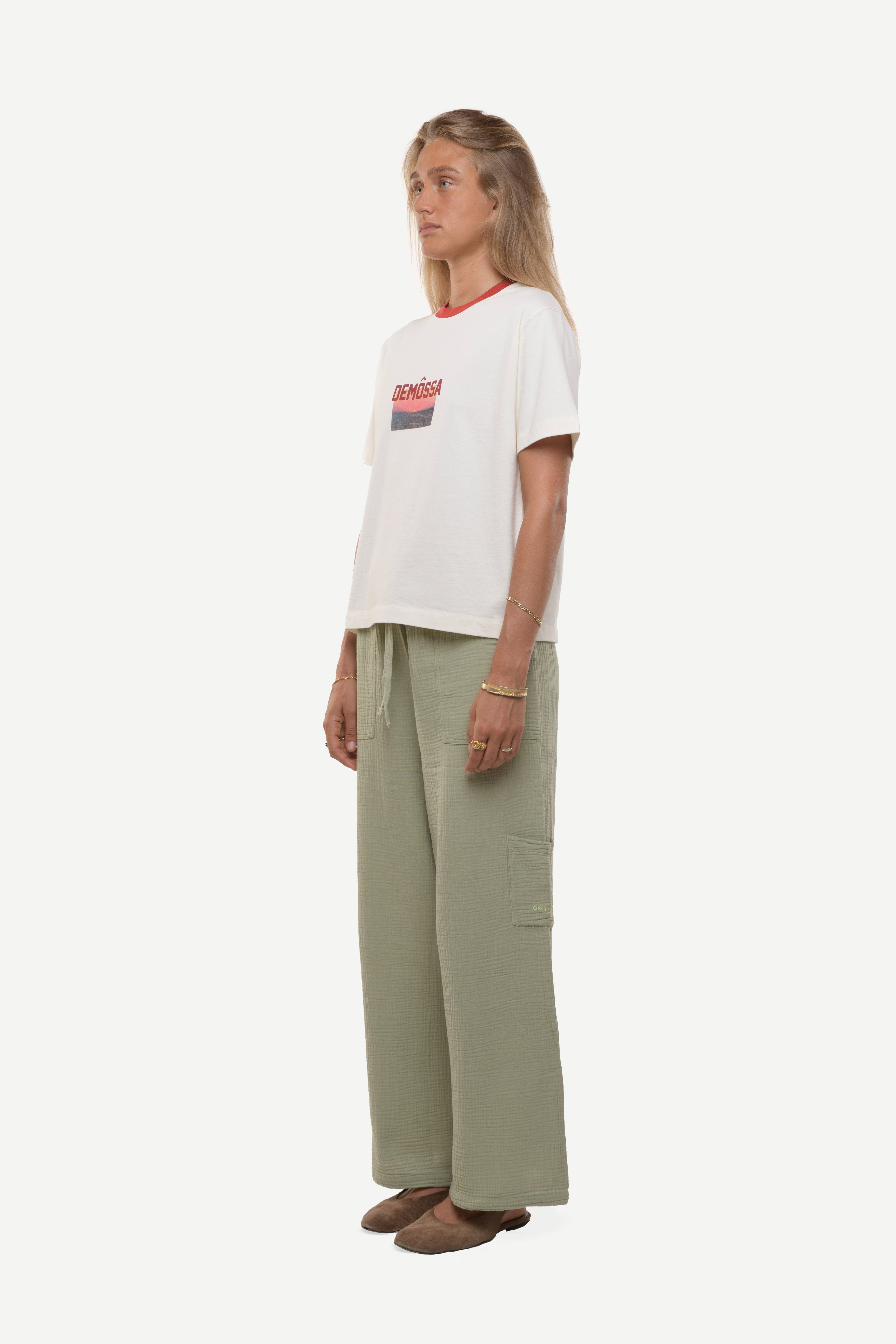 Side view of woman wearing a white t-shirt with red demossa logo and sunset picture with green cotton pants white background