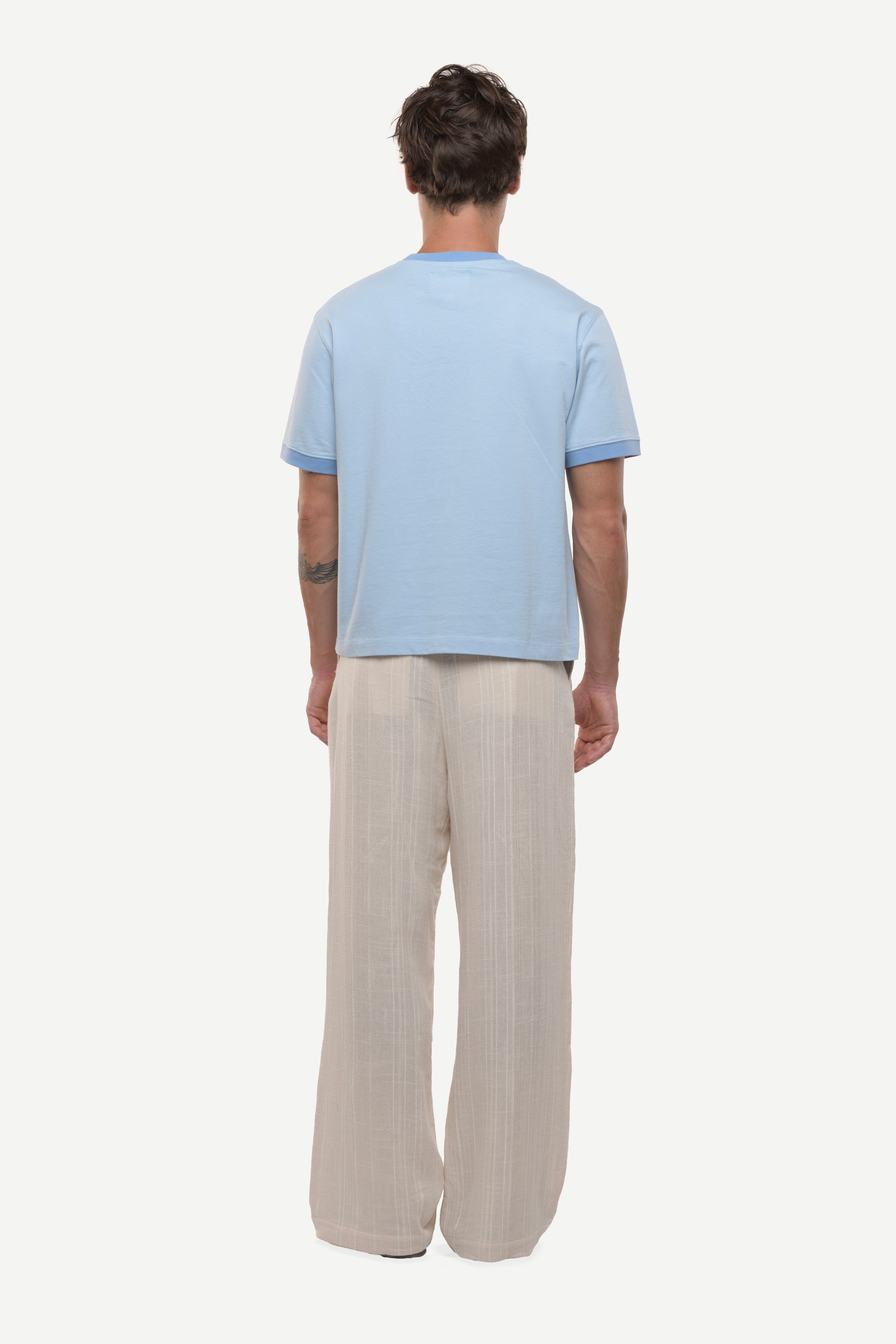 Back view of a man wearing a light blue t-shirt with contras ribs and beige pants on a white background