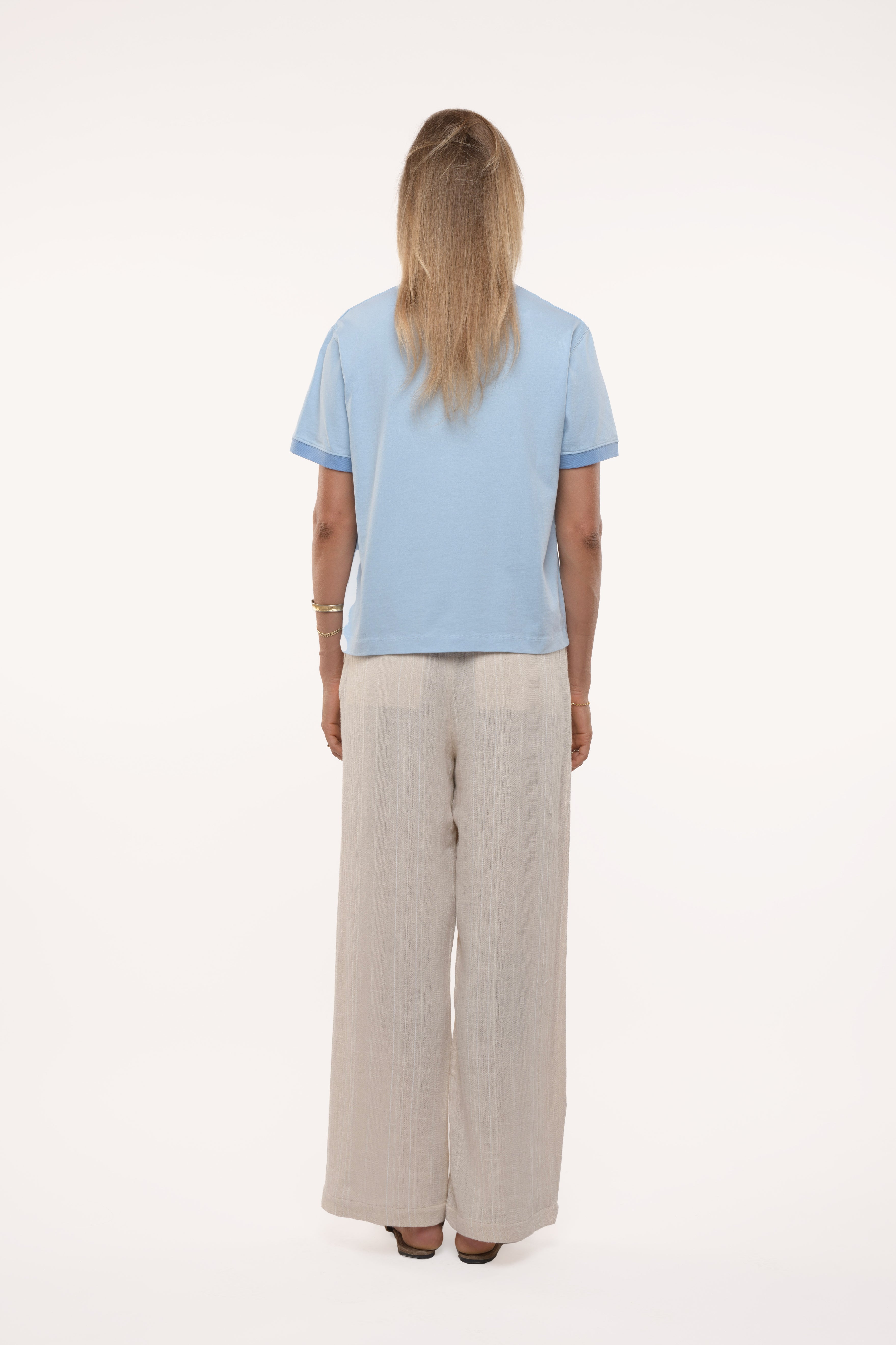 Back view of a woman wearing a light blue top and beige pants on a white background