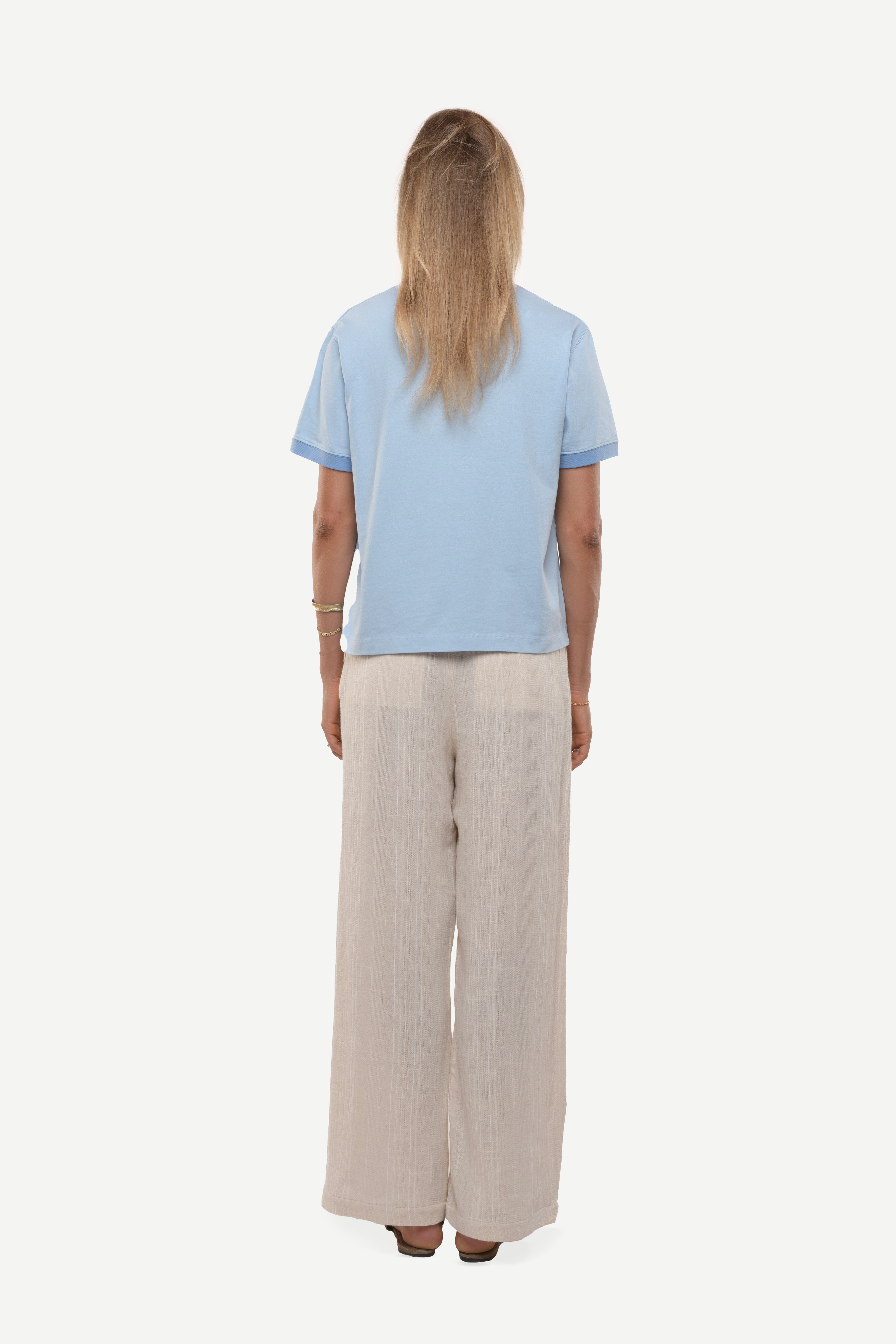 Back view of a woman wearing a light blue t-shirt with contras ribs and beige pants on a white background