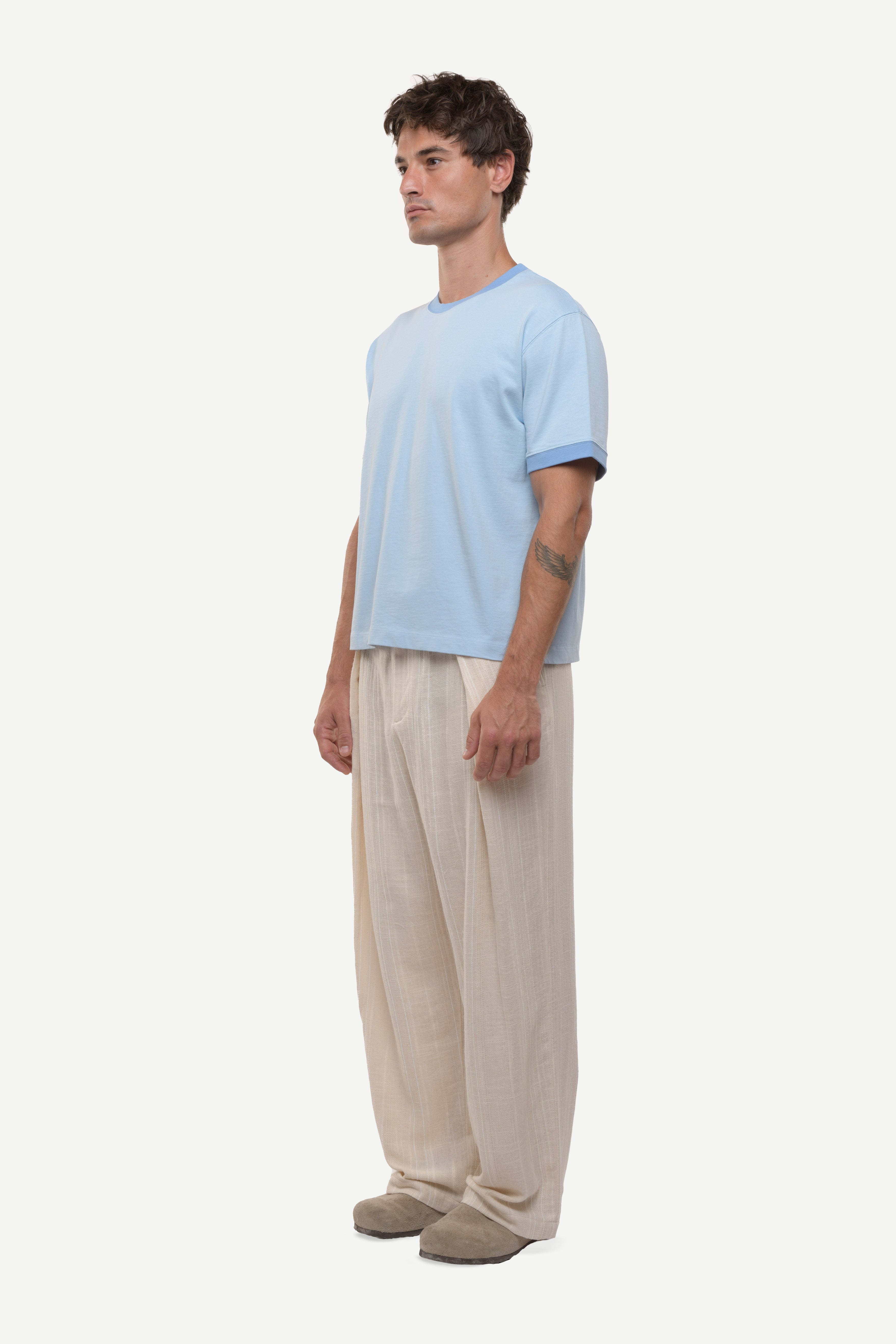 Side view of a man wearing a light blue t-shirt with contras ribs and beige pants on a white background