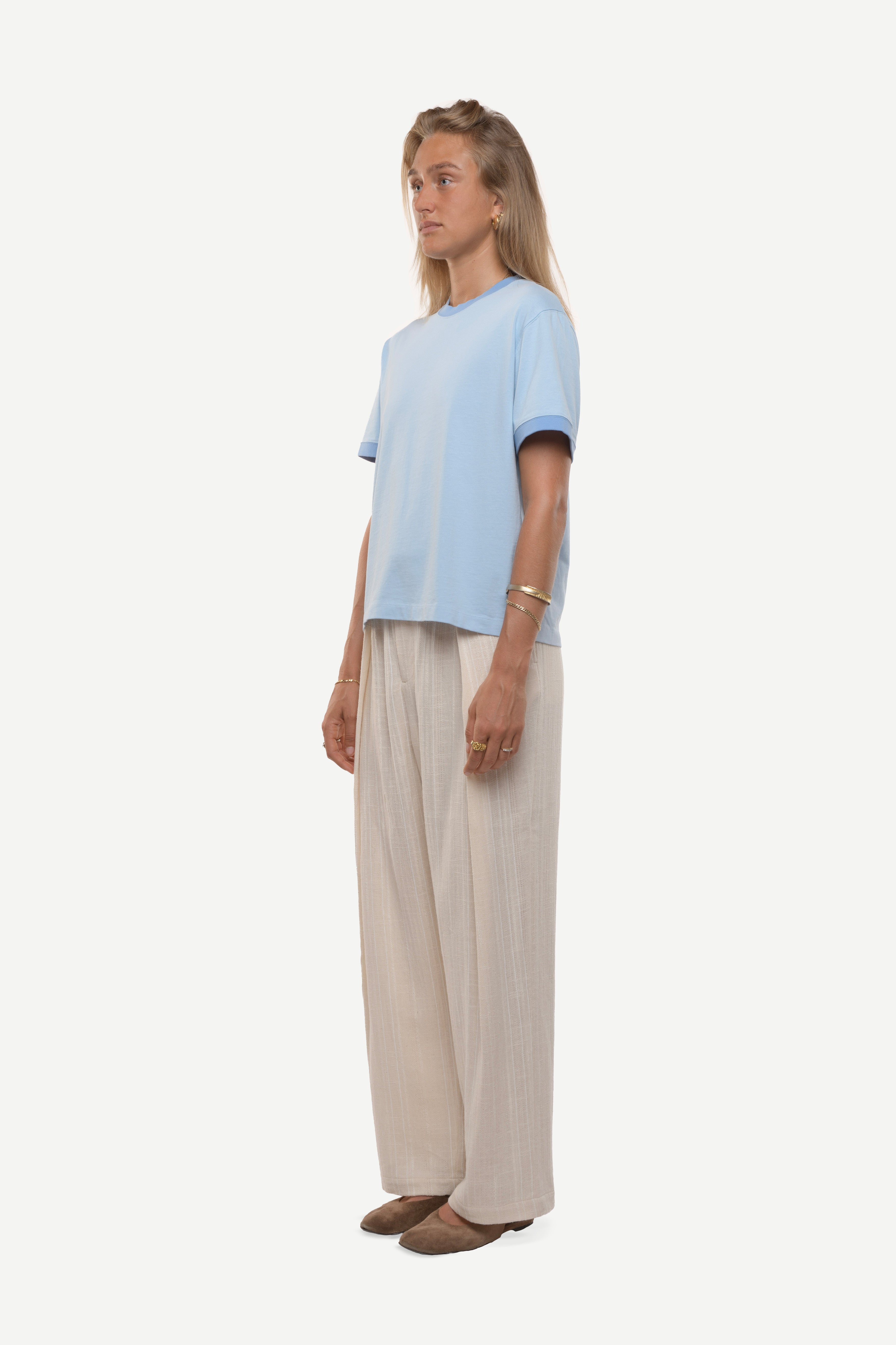 Side view of a woman wearing a light blue t-shirt with contras ribs and beige pants on a white background