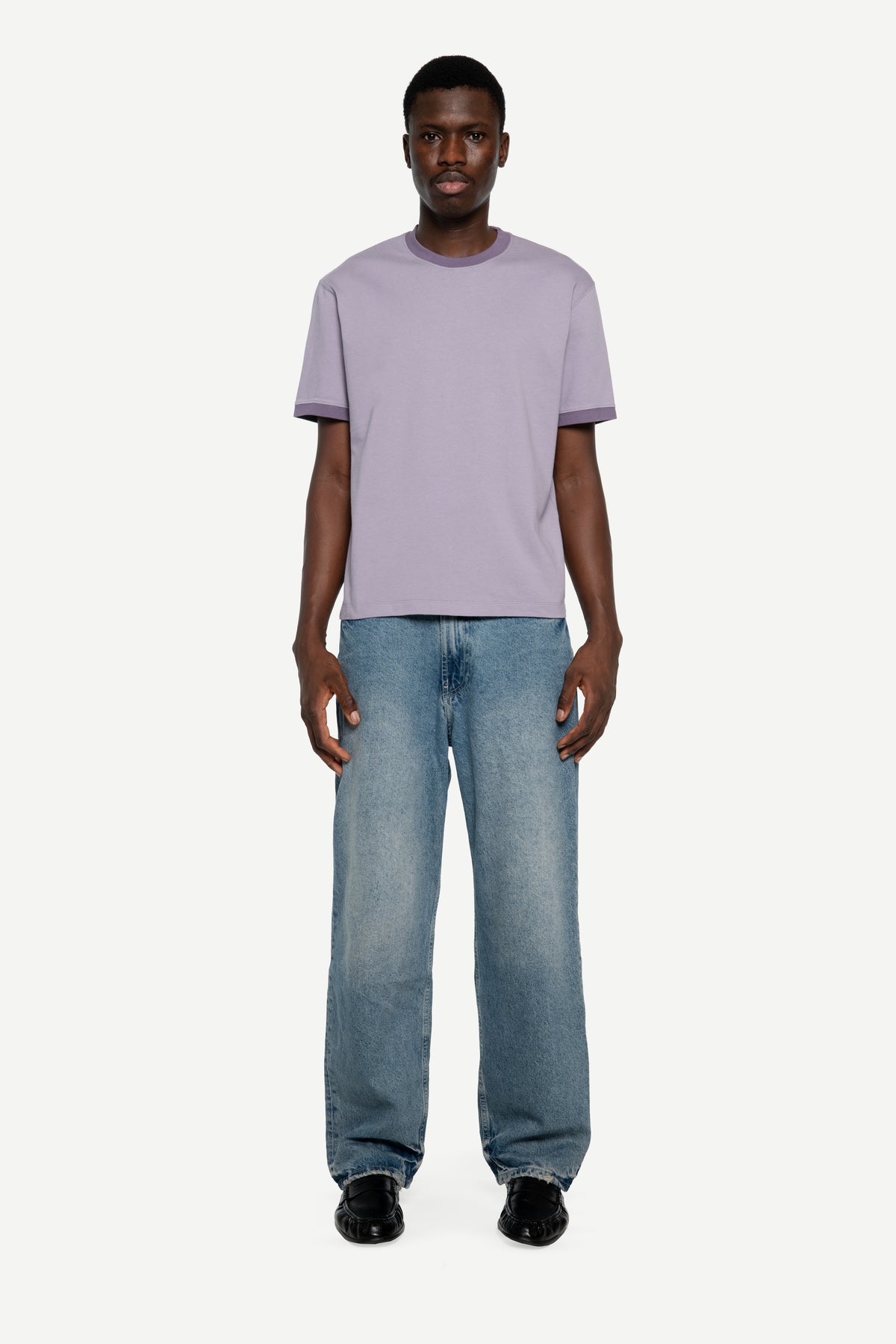 A man modeling a light purple short-sleeve crewneck t-shirt and wide-leg blue jeans against a plain light gray background.