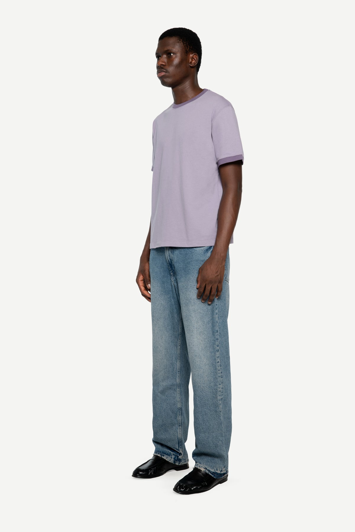 A man wearing a light purple ringer t-shirt with contrasting dark purple cuffs, light blue jeans, and black leather shoes, standing against a plain off-white background.