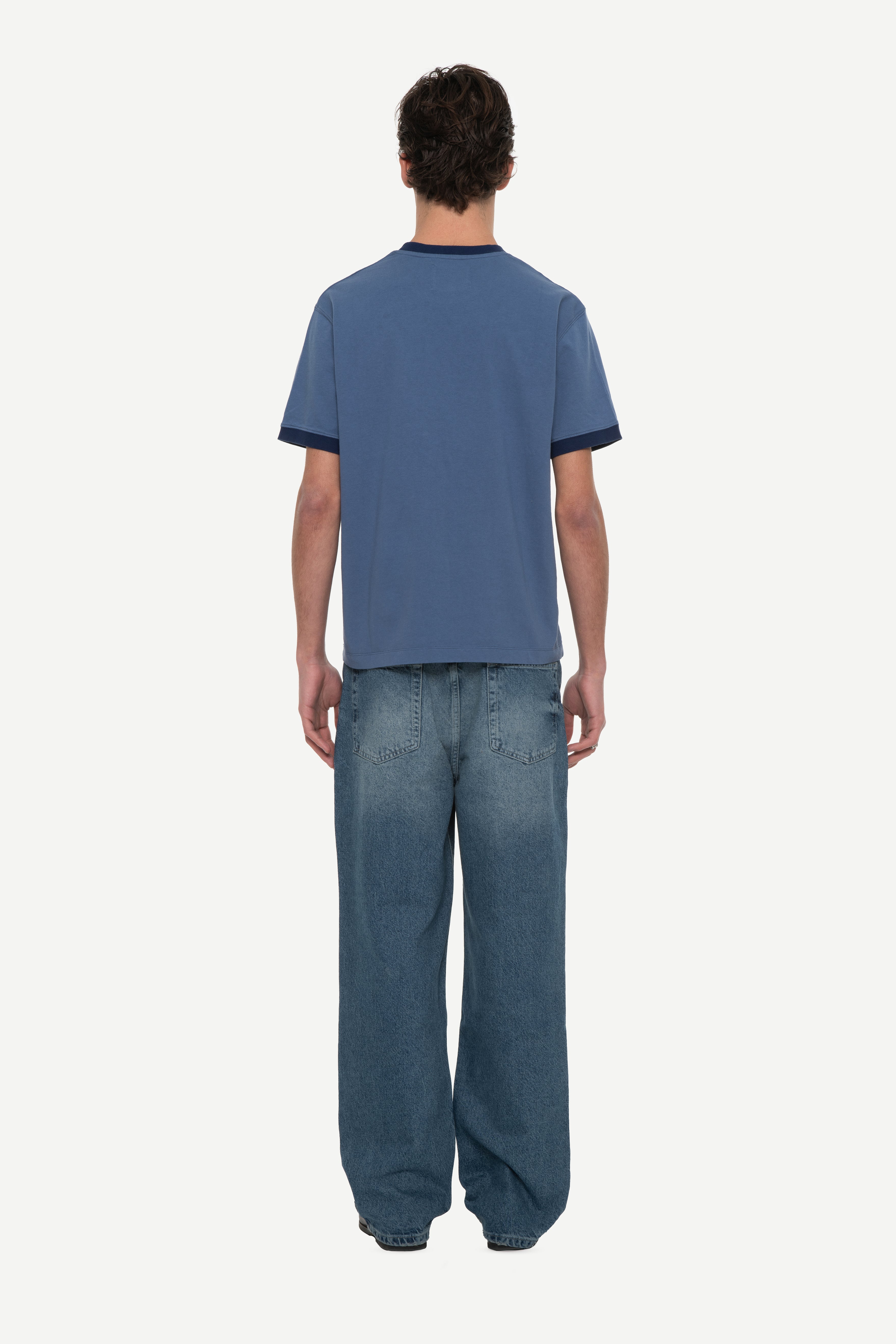 back view of man wearing a navy t-shirt with contrast collar and arm rib and blue jeans on a white background