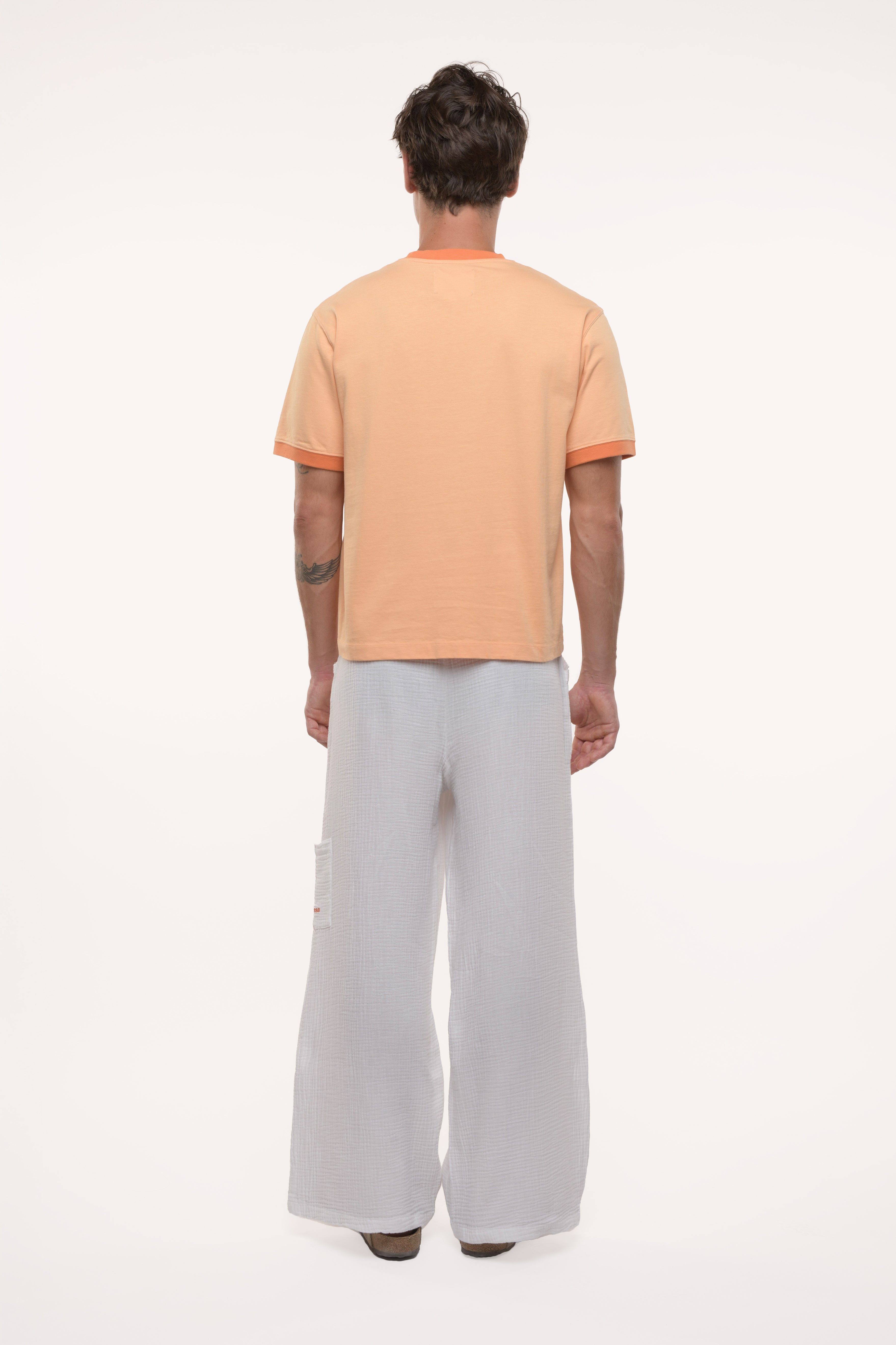 Back view of a man wearing an orange t-shirt with contrast collar and sleeve ribs and light gray pants on a white background