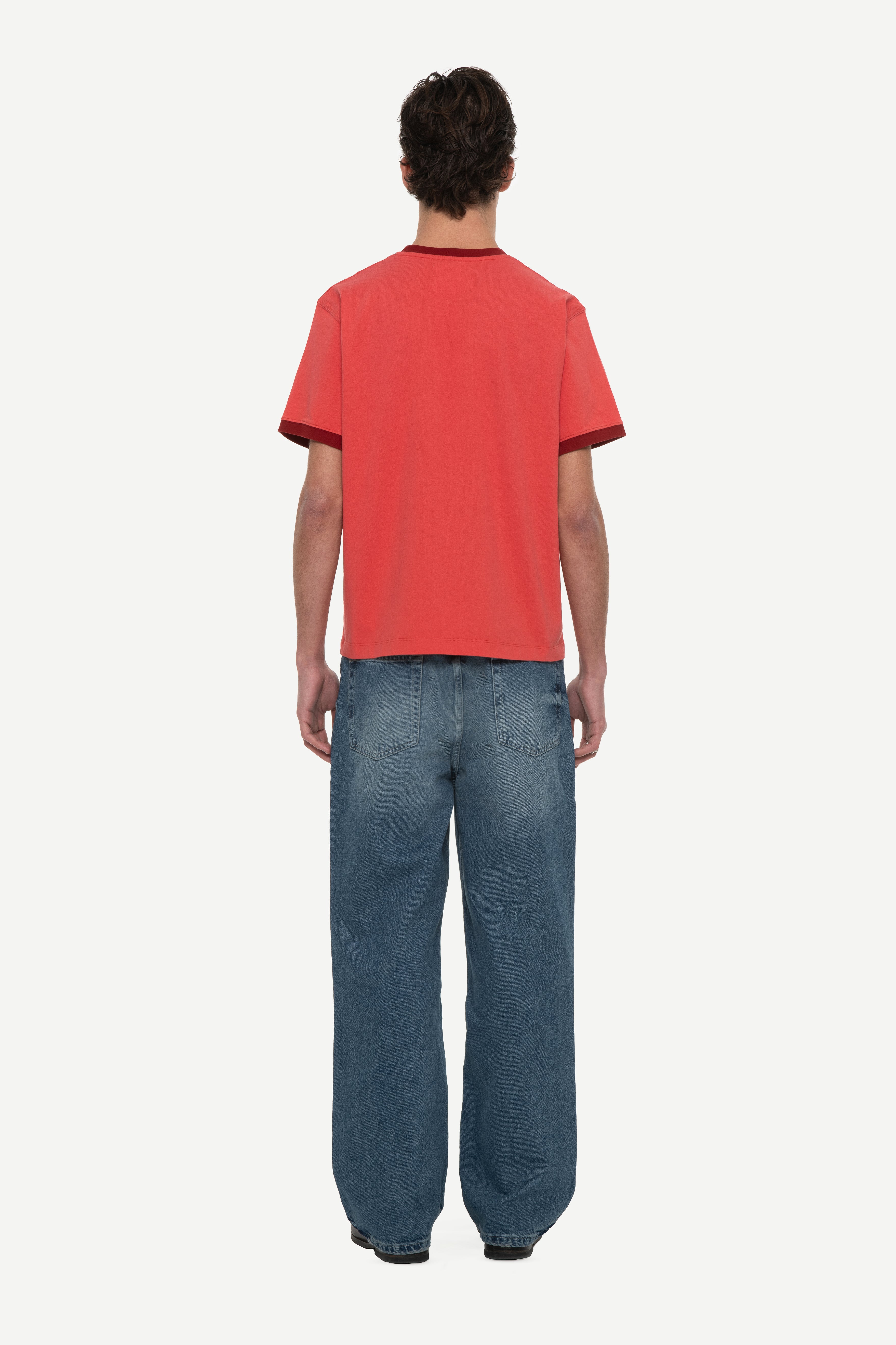 back view of man wearing a red t-shirt with contrast collar and arm rib and blue jeans on a white background