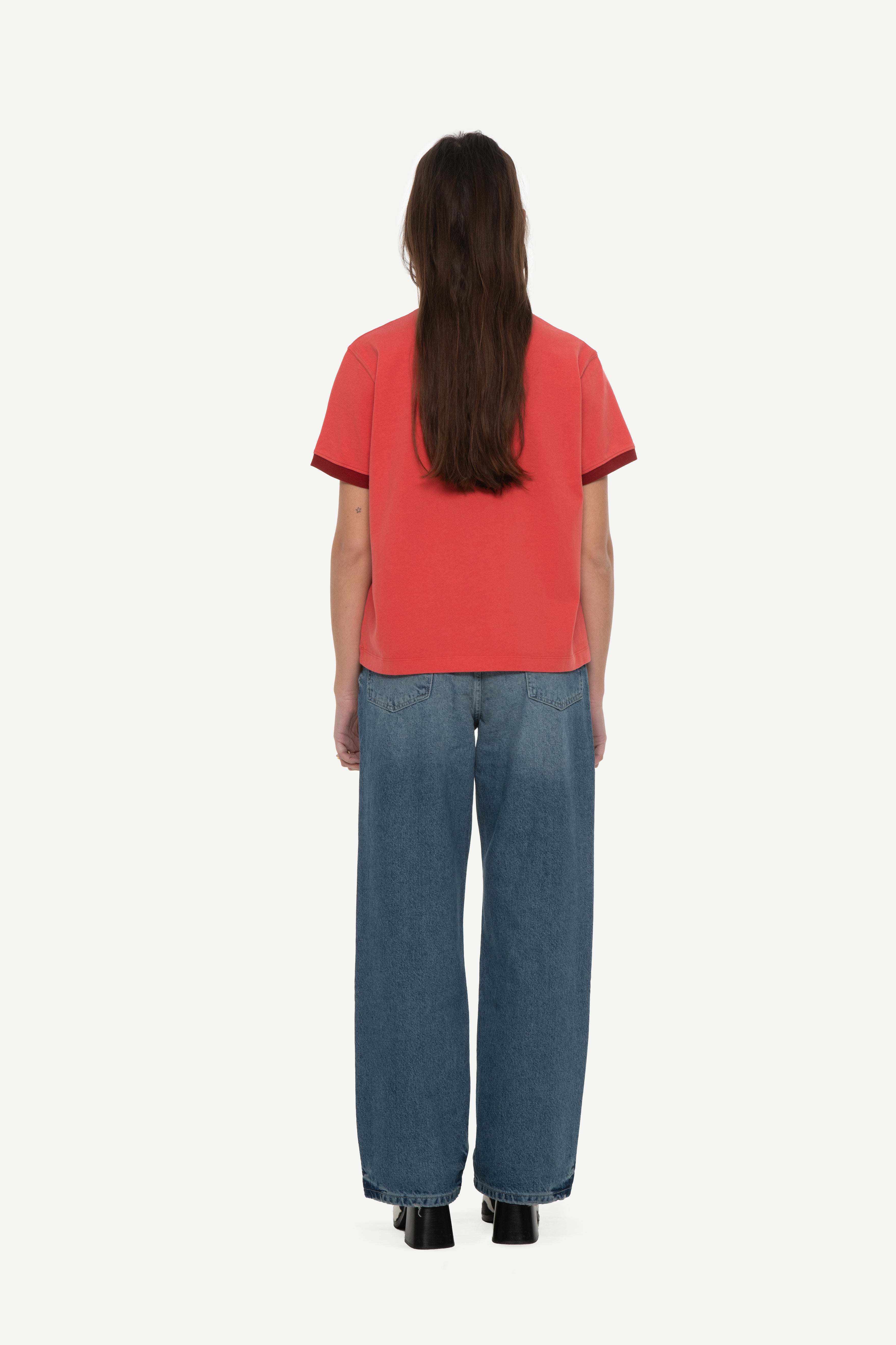 back view of woman wearing a red t-shirt with contrast collar and arm rib and blue jeans on a white background