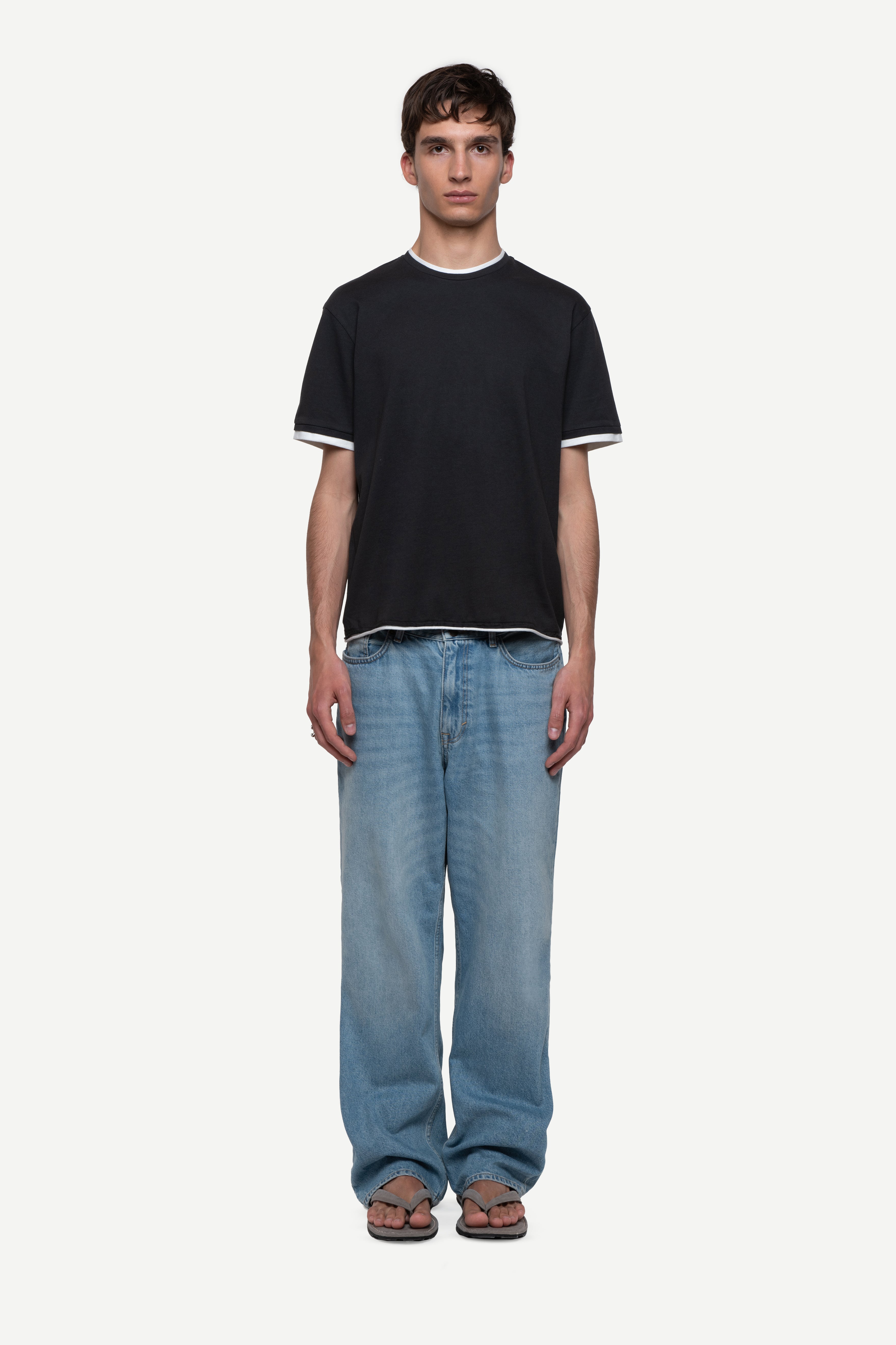 front view of man wearing a regular fit black short-sleeved t-shirt with white double-layered neckline, sleeves and hem, blue jeans and flip flops on a white background