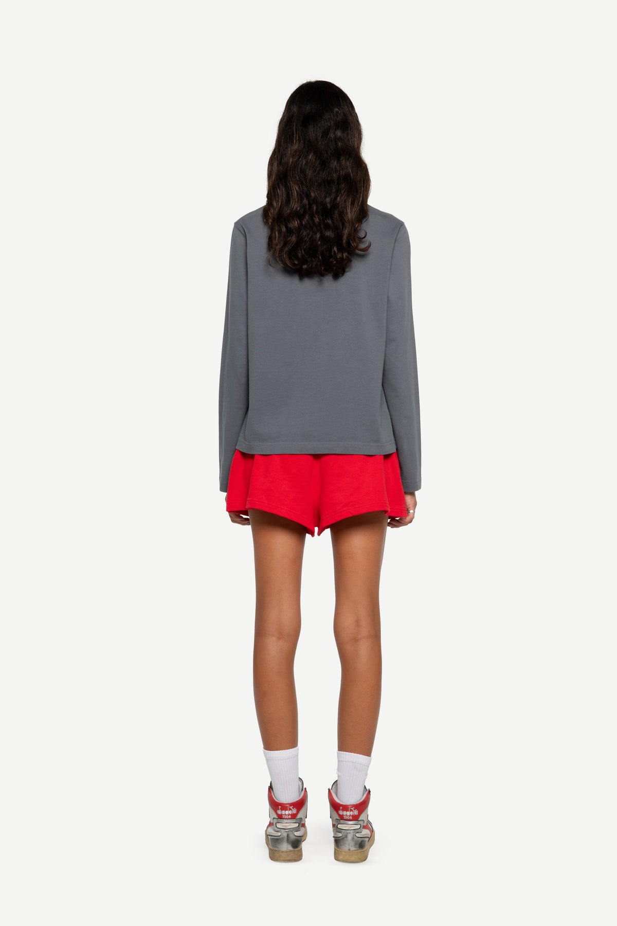 A model shown from the back wearing a gray long-sleeve top, red shorts, white socks, and red-and-gray sneakers against a plain light gray background.