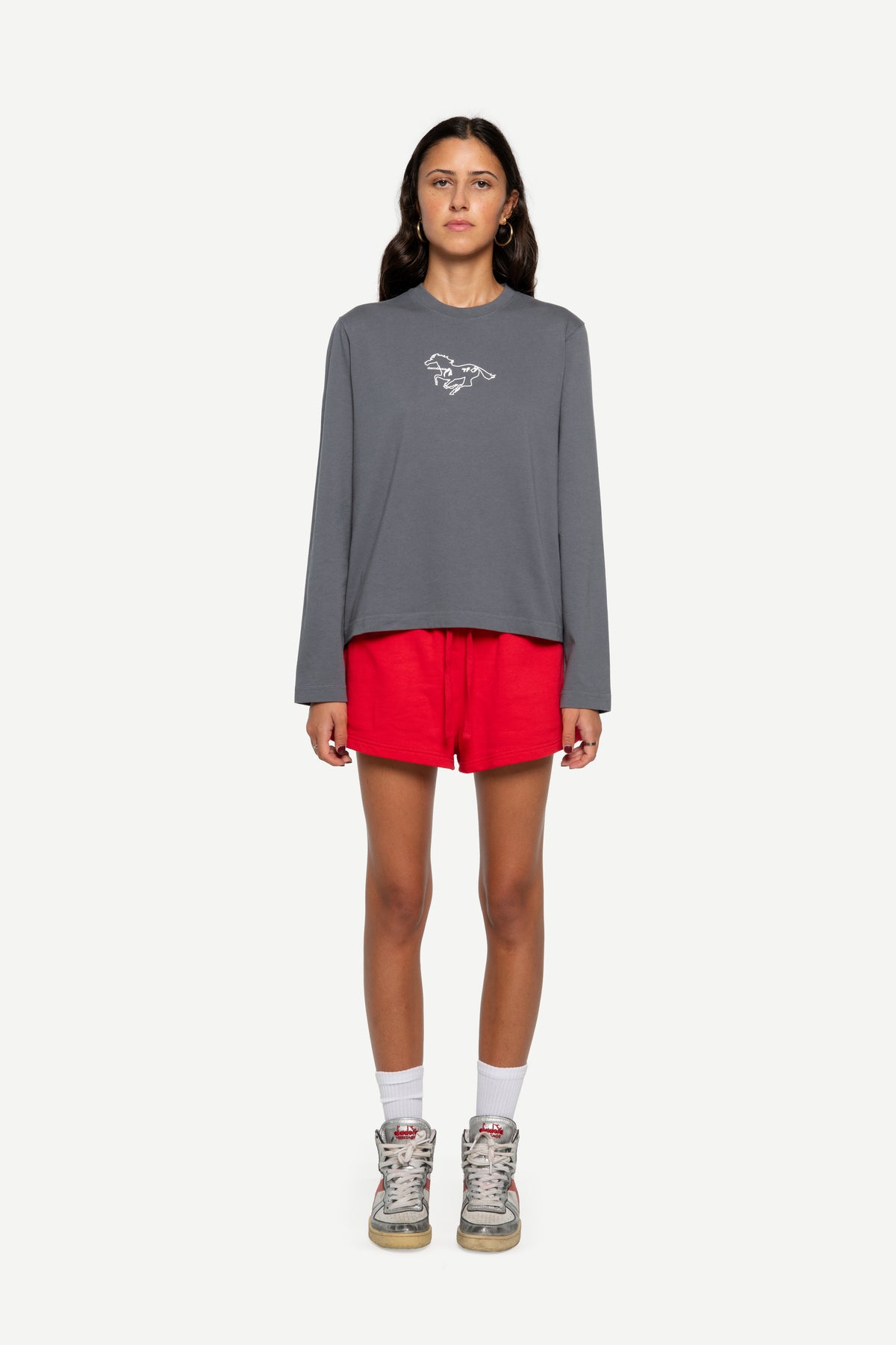 A model stands against a light gray background wearing a dark gray long-sleeve t-shirt with a small white graphic on the chest, paired with bright red athletic shorts, white socks, and gray sneakers.