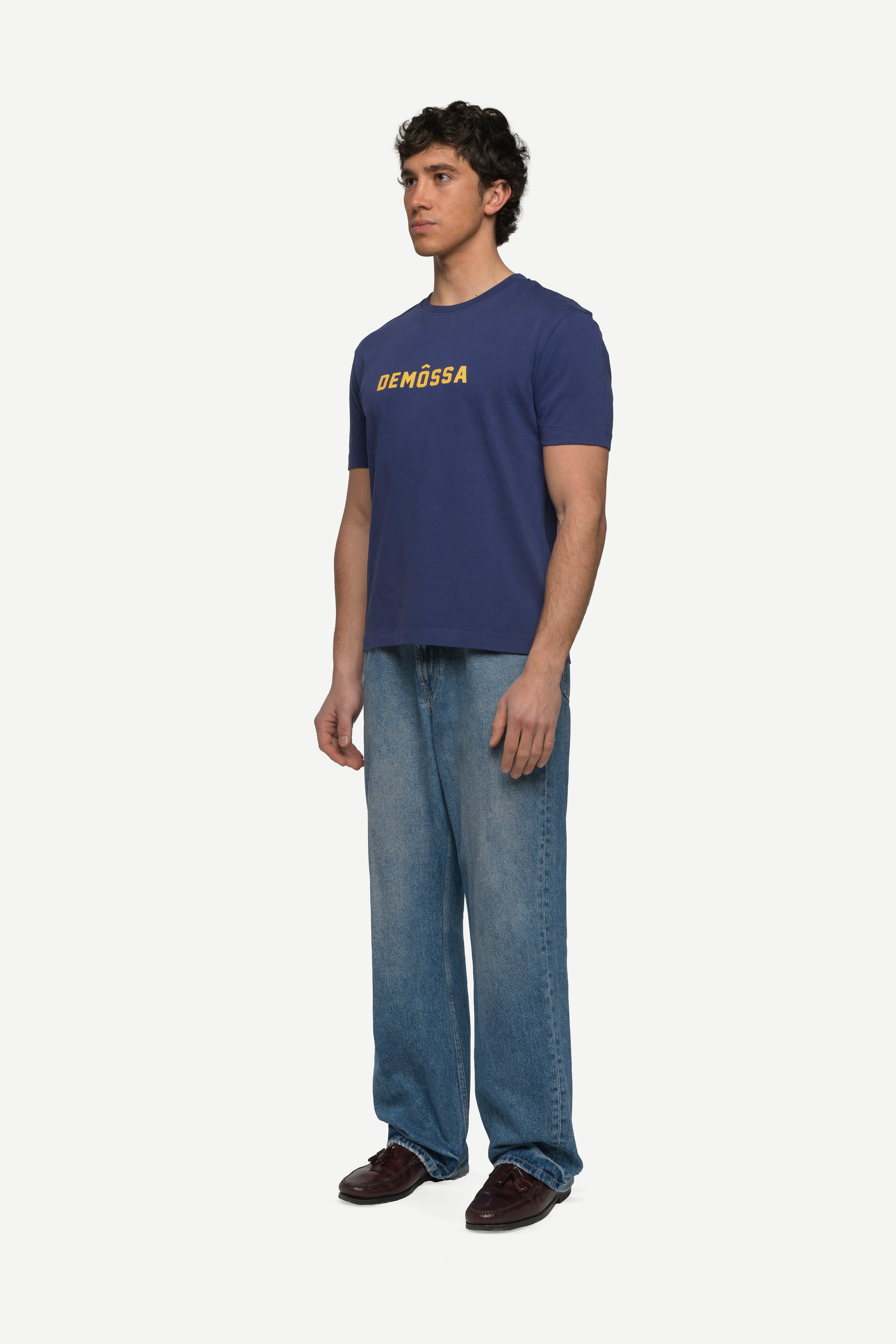 side view of man wearing a navy t-shirt with yellow demossa logo on the chest and blue jeans on a white background