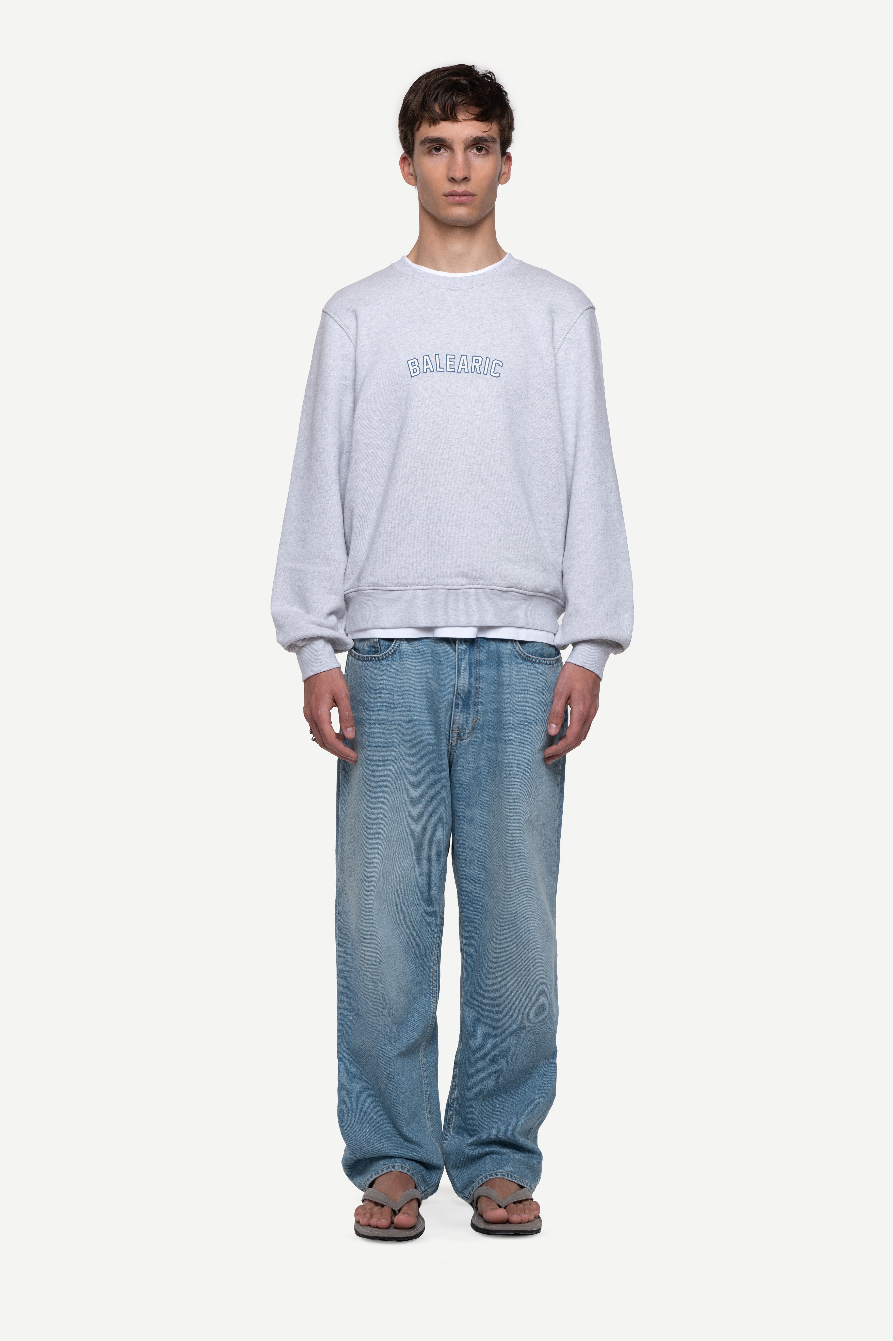 front view of man wearing a melange grey relaxed-fit sweatshirt with "Balearic" print on the chest, blue jeans and flip flops on a white background