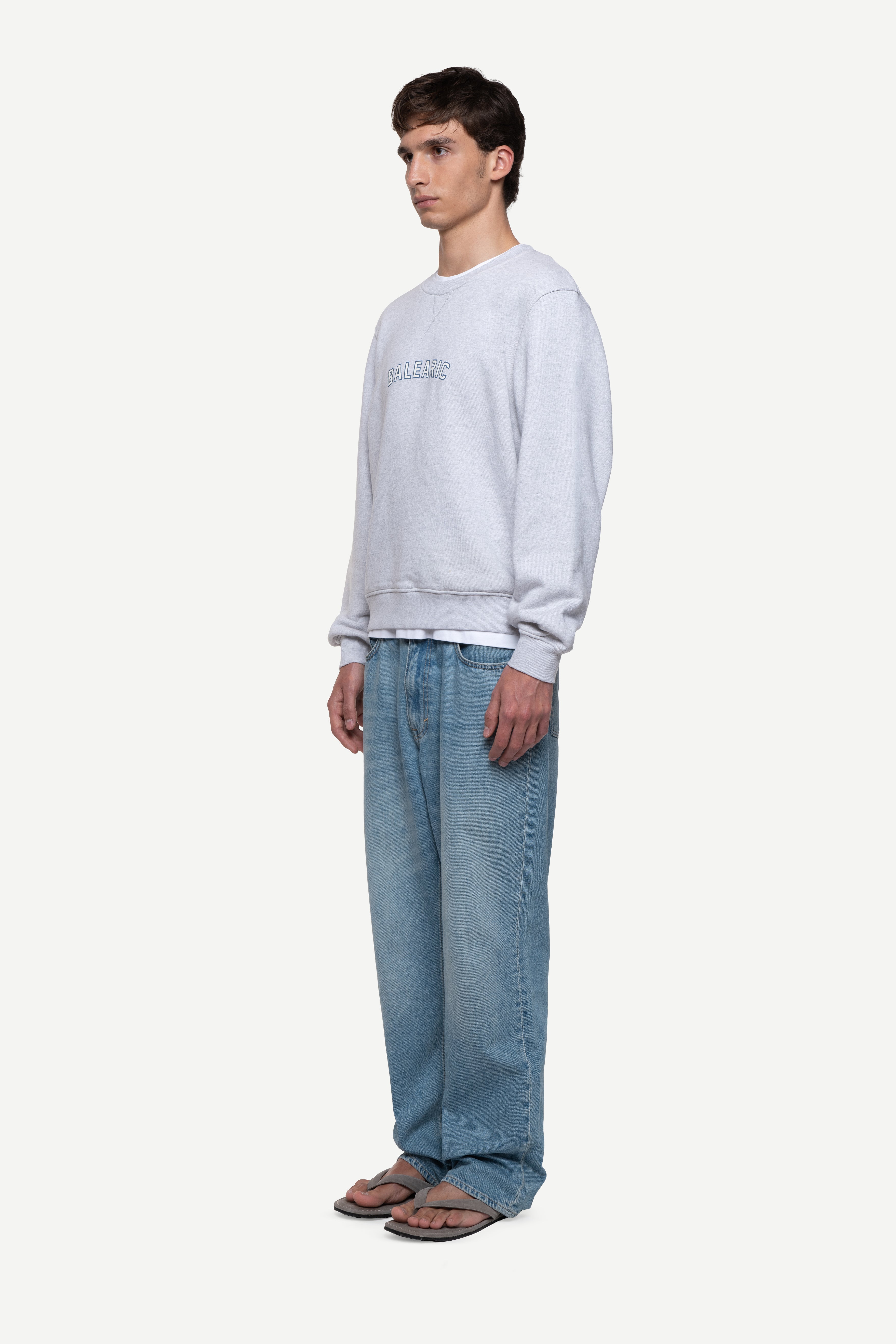side view of man wearing a melange grey relaxed-fit sweatshirt with "Balearic" print on the chest, blue jeans and flip flops on a white background