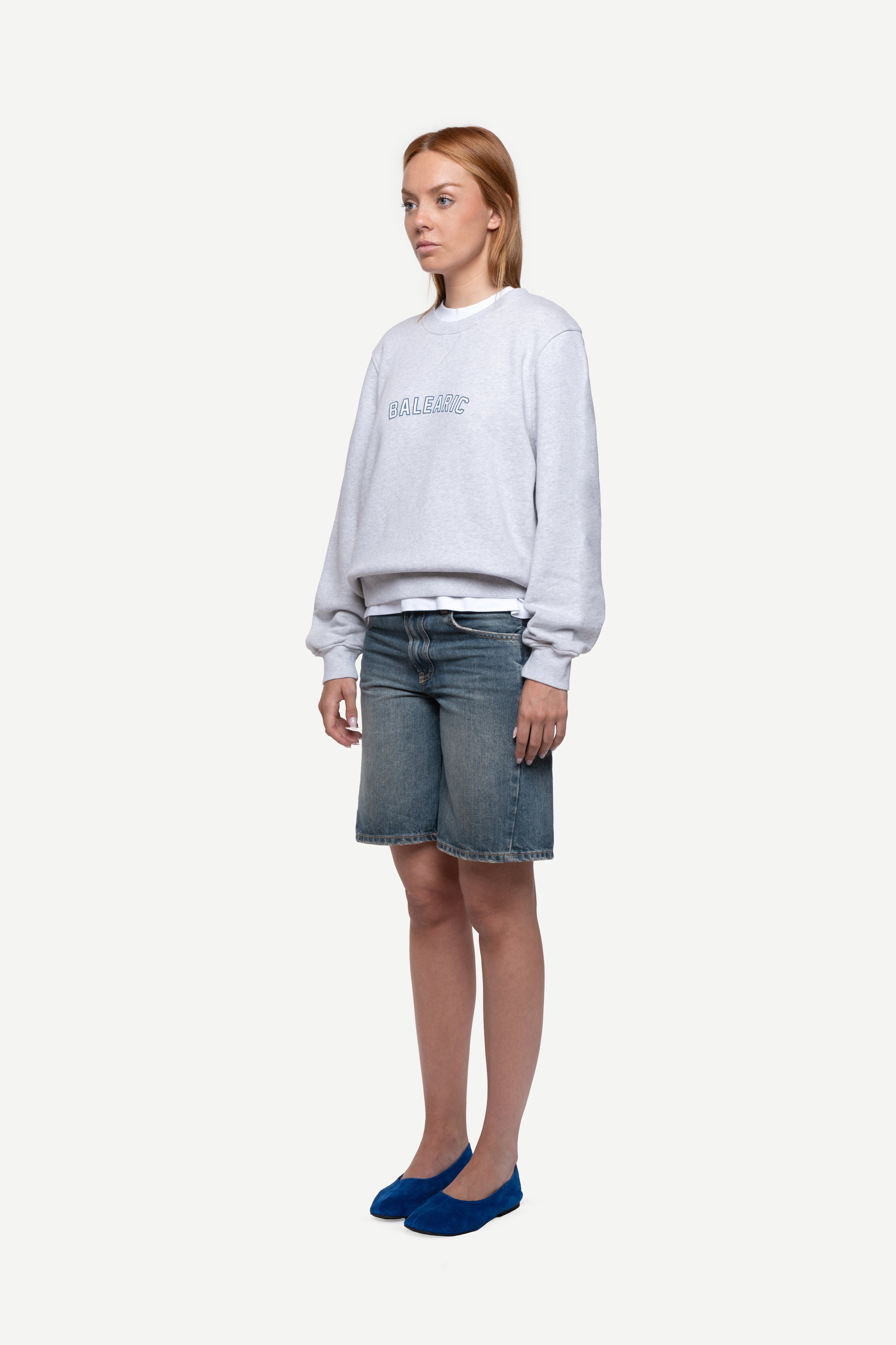 side view of woman wearing a melange grey relaxed-fit sweatshirt with "Balearic" print on the chest, blue short jeans and ballet flats on a white background